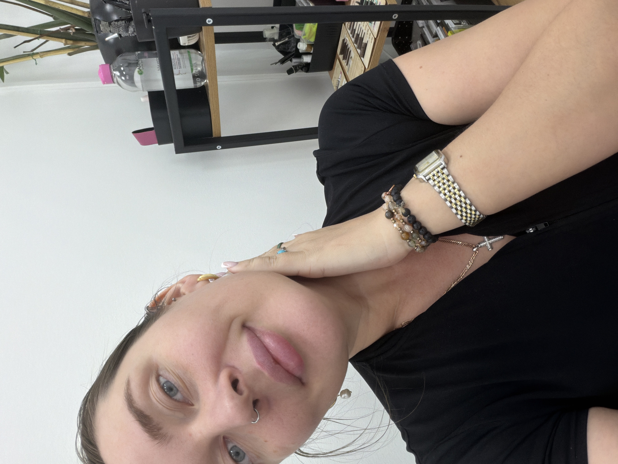 A selfie of a young adult female with fair skin and light blue eyes is visible, taken indoors. She has pierced ears and a nose ring and is wearing a black top. She is wearing a watch and bracelets on her left wrist, and a necklace with a cross pendant. The background features a white wall and a black metal shelf with various items, including a bottle with a pink cap and other beauty products. The location is not identifiable as Moscow, Russia.