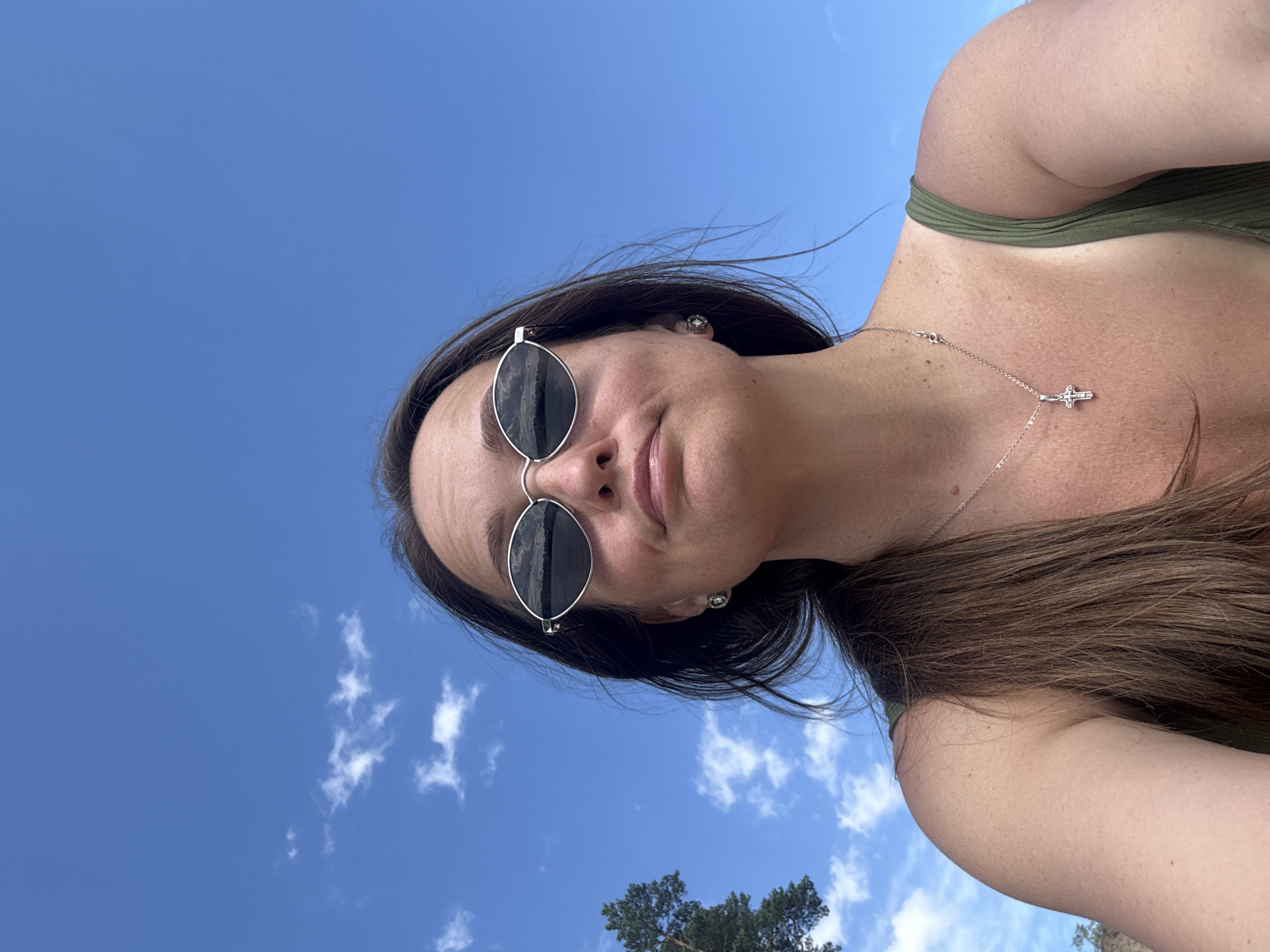 A selfie shows a woman with dark hair and sunglasses against a bright blue sky with scattered white clouds. She is wearing a green top and a silver necklace with a cross pendant. To the left of the woman, a portion of a pine tree is visible, suggesting an outdoor setting, possibly near a body of water given the reflection in her sunglasses. The image was taken on a sunny day. The location is identified as Novyi, Russia.