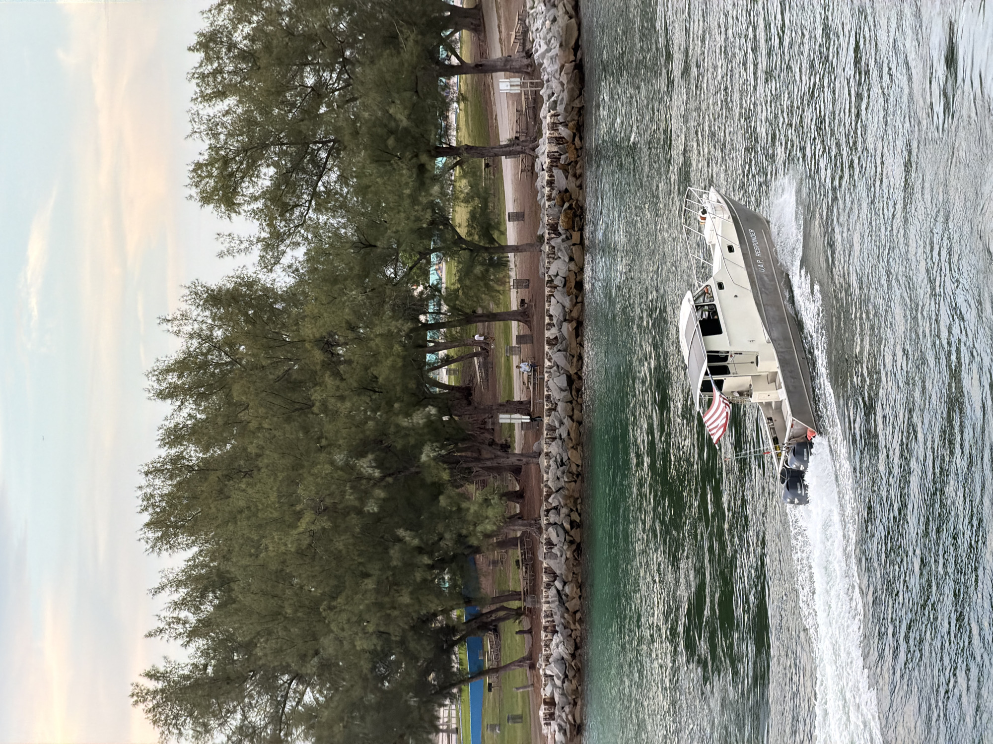 A medium shot captures a boat with "UAP RESPONDER" written on its side, moving through green-tinted water, leaving a white wake. The boat, with twin outboard motors, displays an American flag. In the background, a rocky shore separates the water from a park area with picnic tables, trees, and a few people. The sky above is a pale gradient of pink and blue, with a faint distant airplane. The scene appears to be a coastal area in Bal Harbour Village, Florida, United States of America.
