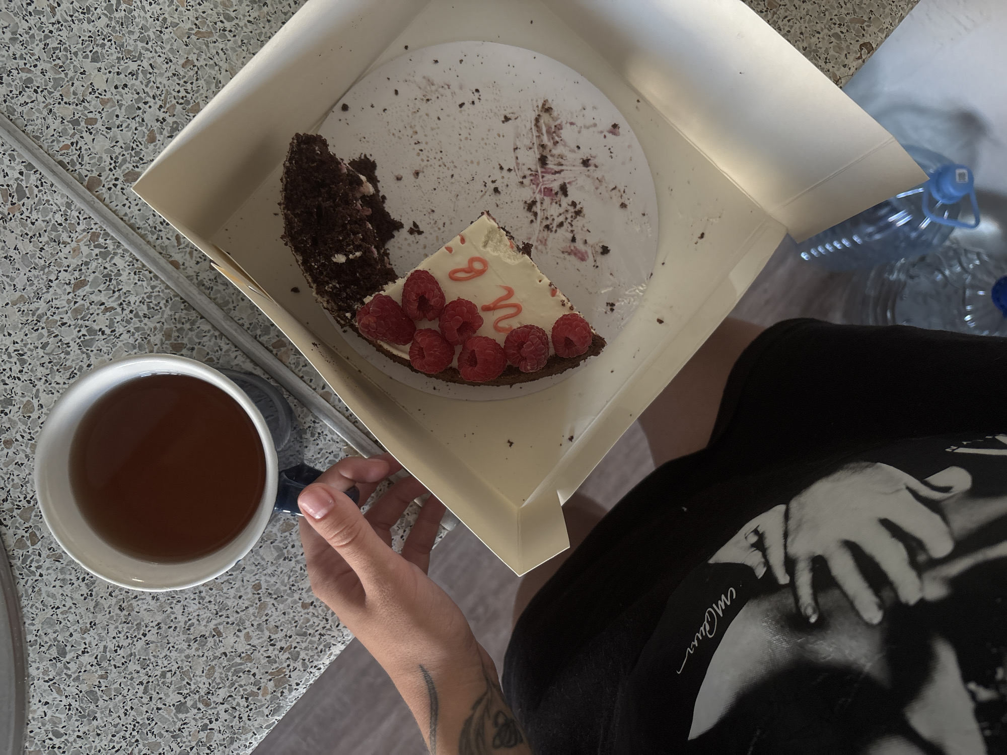 In Rostov-on-Don, Russia, a person is holding a mug of tea next to a box containing a half-eaten slice of chocolate cake decorated with raspberries and pink frosting. The cake has the Cyrillic letters "и в" written on it. Crumbs and frosting are scattered on the cake's plate and in the box. The person is wearing a black t-shirt with a black and white image on it. A partially visible plastic bottle of water is to the right of the box. The scene is set on a speckled countertop.