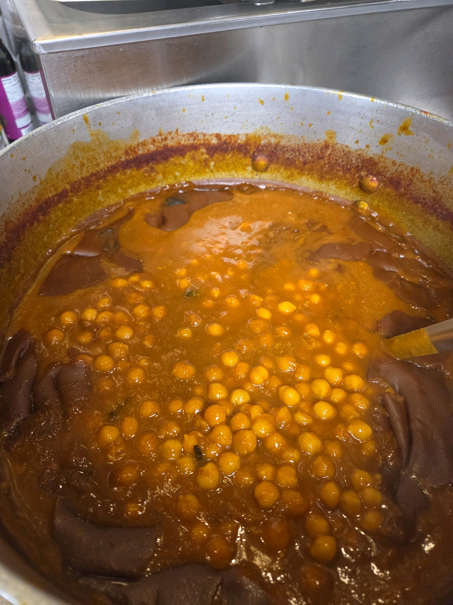 This is a close-up, overhead shot of a large pot filled with a thick, orange-hued stew. The stew contains numerous small, round, yellow chickpeas and appears to have some darker, amorphous ingredients submerged within it. The pot itself is made of a dull metal, and the rim shows some staining from previous use. A portion of a serving utensil, possibly a ladle, is visible on the right side, partially submerged in the stew. The background is out of focus but suggests a commercial kitchen environment with stainless steel surfaces and some bottles or containers. The image appears to be taken in Haddonfield, United States of America.