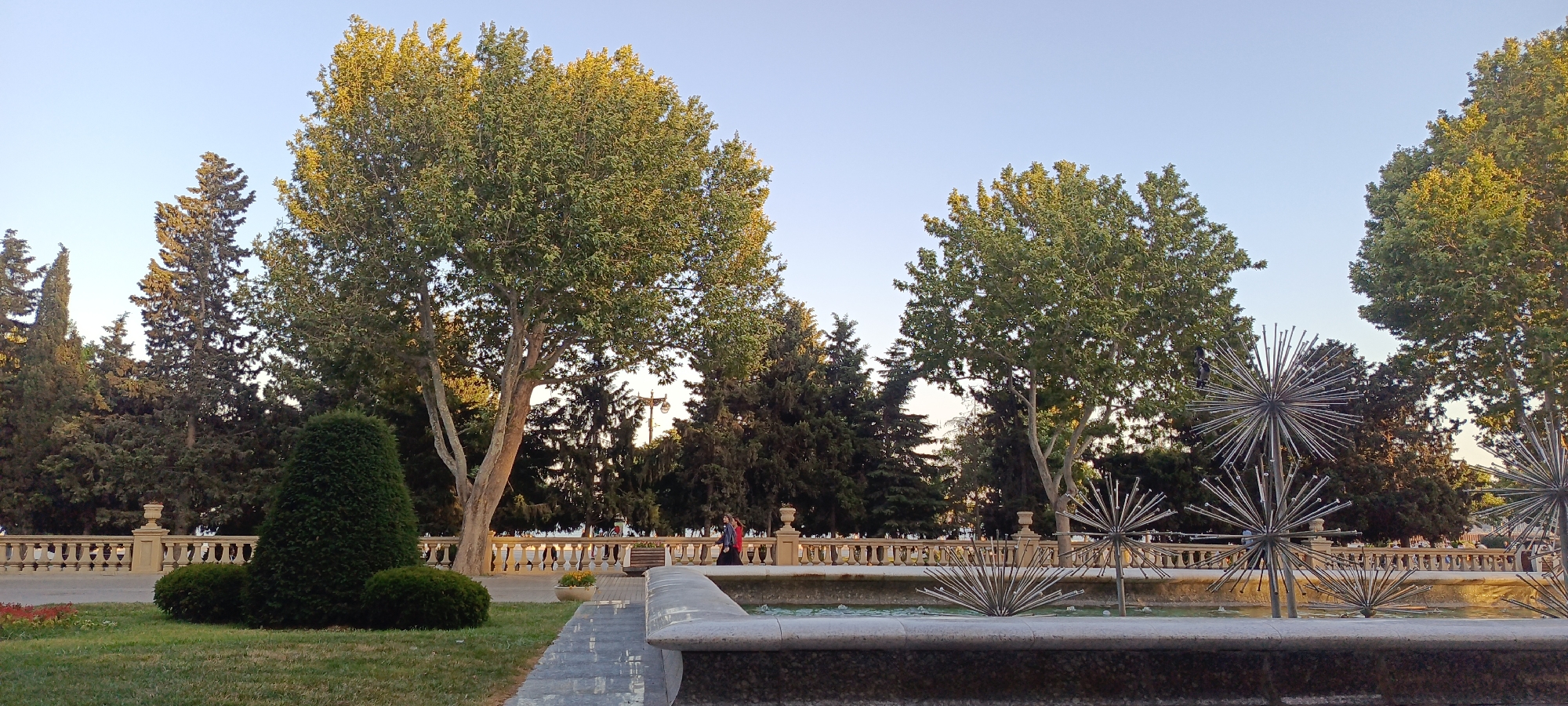 This is a wide-angle photograph taken in Sabail Raion, Azerbaijan, likely in a park setting near the Caspian Sea. The image captures a sunny day with a clear, pale blue sky. In the foreground, a stone fountain basin with dark, speckled stone detailing is visible. Within the basin, several metallic, spiky, spherical sculptures, resembling sea urchins, are submerged in shallow water.
To the left of the fountain, a neatly trimmed, conical green hedge stands prominently on a grassy area. Behind it, more manicured bushes are visible. A grey paved walkway, bordered by grass, leads into the scene from the lower left.
A low, ornate stone railing with decorative balusters runs across the middle ground of the image, separating the fountain area from a backdrop of trees and a walkway. Two individuals, one wearing a red top and the other in blue, are walking along this path, partially obscured by the railing.
The background is filled with a dense arrangement of various trees. Tall, slender evergreen trees with dark green foliage are interspersed with broader deciduous trees featuring lighter green leaves, some of which are tinged with yellow from the sunlight. In the distance, beyond the trees and railing, a hint of the Caspian Sea or a large body of water is visible, suggesting a coastal location. The overall impression is one of a well-maintained public space designed for leisure and enjoyment.