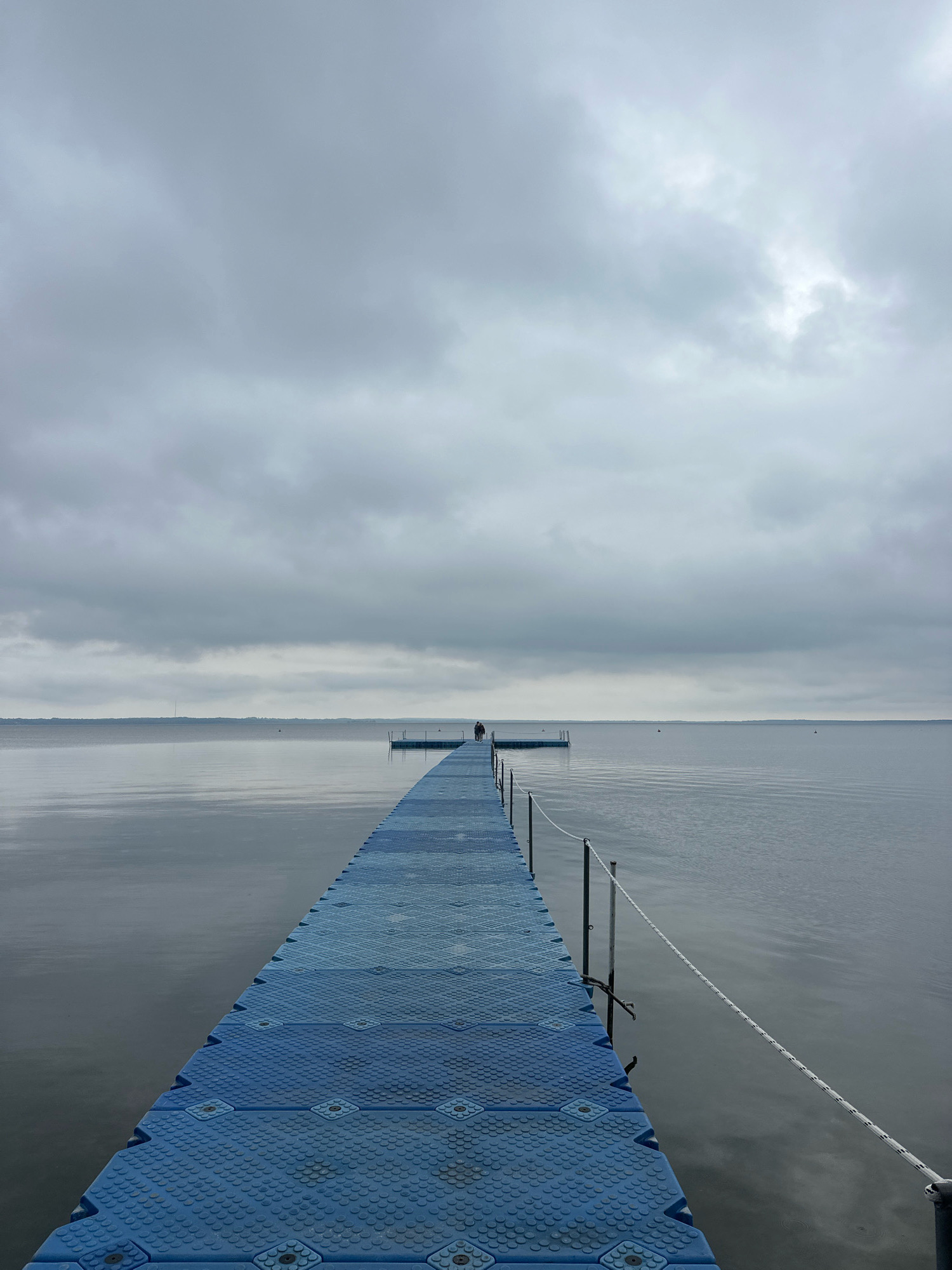 The image shows a long, blue modular pontoon walkway extending over a calm, gray body of water. The sky is overcast with heavy, textured gray clouds. In the distance, on the horizon, a faint landmass is visible. Near the end of the pontoon walkway, a single person stands, facing away from the camera. Two smaller buoys are visible in the water on the left side of the pontoon. A white rope railing runs along the right side of the walkway. The scene appears to be located in the Narach Rural Council, Belarus, given the characteristic landscape and overcast weather conditions common to the region, particularly during transitional seasons.