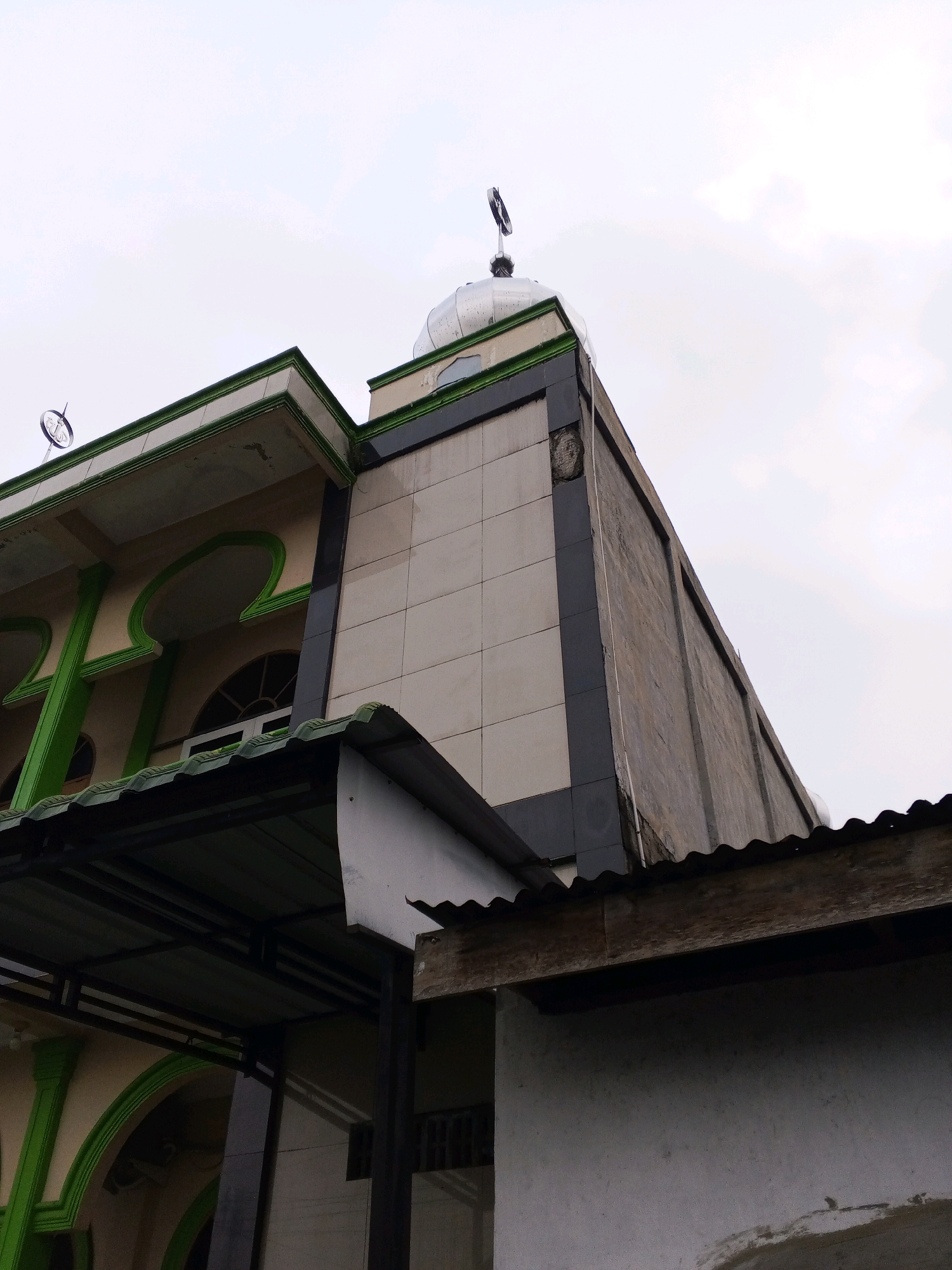 An angled outdoor, low-angle shot captures the upper section of a mosque in Lama, Indonesia. The building features a dome topped with a crescent moon symbol, a tiled facade, and decorative green arches. A metal roof structure extends from the building, and a rough, white-walled structure is visible in the foreground. The sky above is overcast and pale.