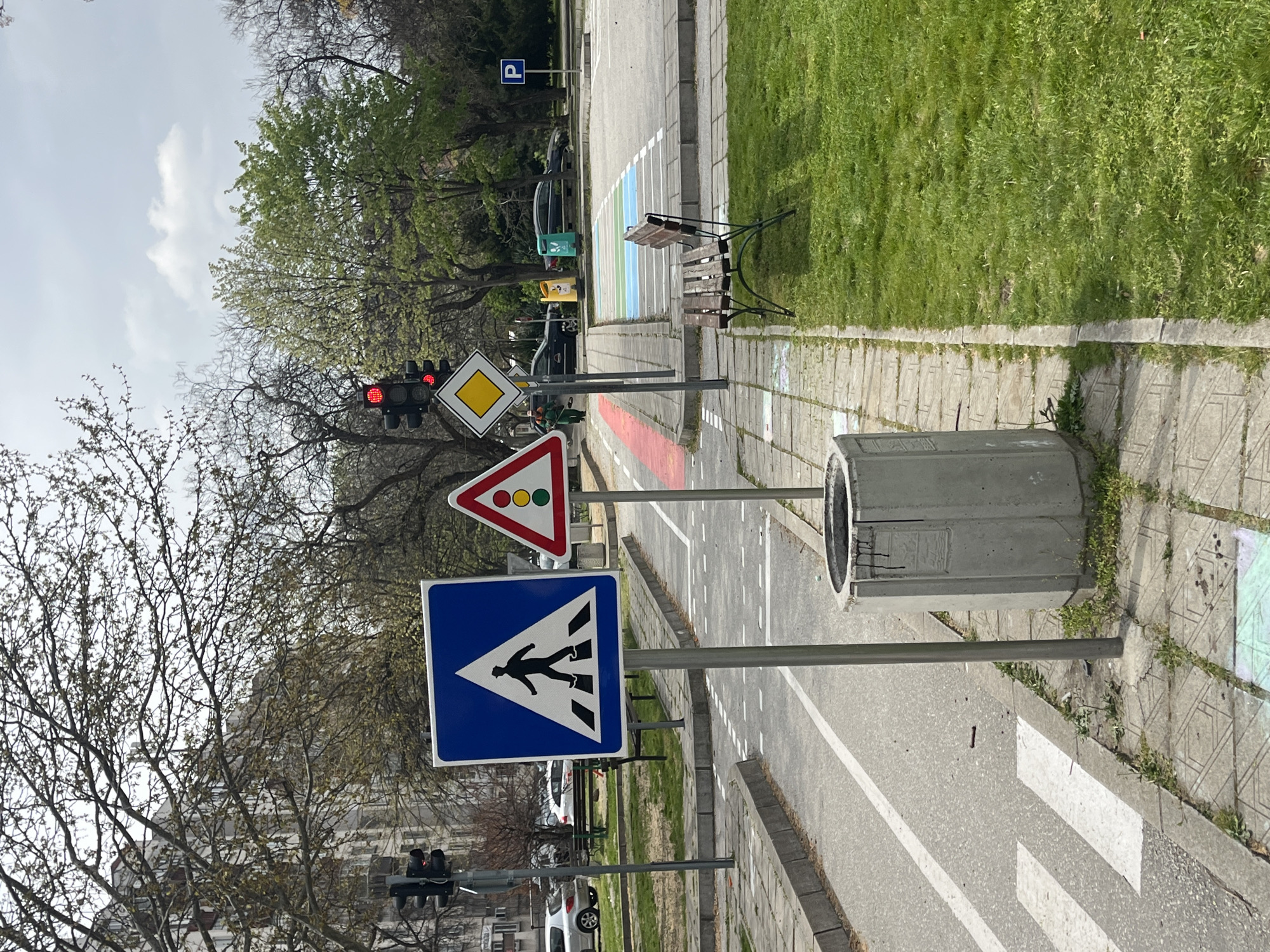 The image depicts a section of a street designed for traffic education in the city of Plovdiv, Bulgaria. The street is lined with sidewalks and has various traffic signs posted along the side. A pedestrian crossing sign, a traffic light signal sign, a priority road sign, and a traffic light are visible. The traffic light is currently showing a red signal. A bench is positioned beside the street, along with a concrete trash can. Grass and trees are present in the background. Cars are parked on the side of the street further in the background. A parking sign is also visible. The sky is overcast.