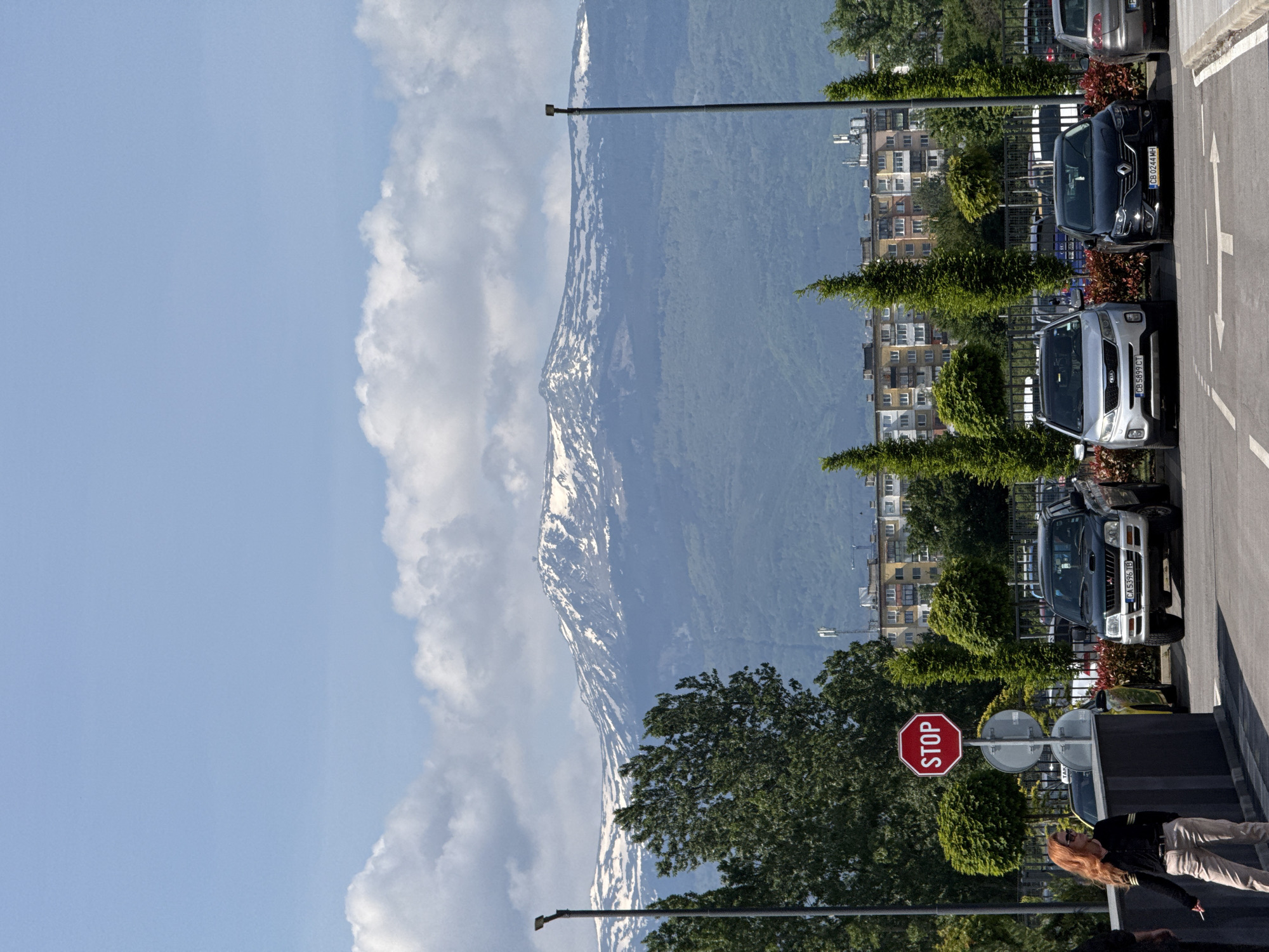 The image shows an outdoor scene in Sofia, Bulgaria, likely near a parking area on the edge of the city with a mountain backdrop. The sky is light blue with large white clouds. The mountain, likely Vitosha, is partially covered in snow. In the midground are buildings of varying heights, suggesting a mixed residential and commercial area. A row of trees lines the side of a road or parking lot. Several parked cars of different makes and models are visible. A red and white octagonal "STOP" sign is prominent near the right edge. At the bottom right, a person wearing light-colored pants and a dark jacket is partially visible. The ground is paved. Light posts are also visible.