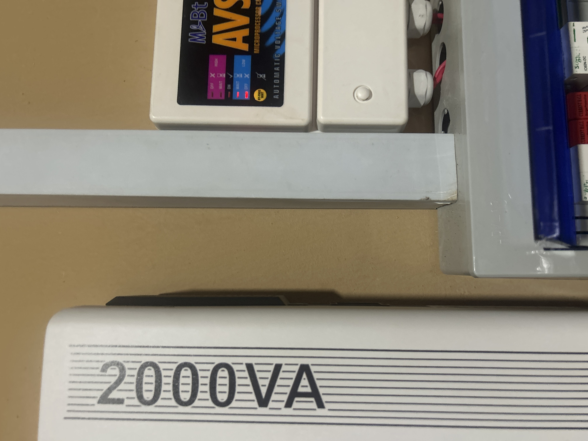 The image shows a close-up of electrical equipment mounted on a wall. On the left is a white vertical unit with the text "2000VA" printed in black. To the right of this unit is a light gray vertical support. Further to the right, a white rectangular device labeled "M & Bt AVS" is visible, with various indicator lights and text such as "MICROPROCESSOR CON" and "AUTOMATIC VOLTAGE SW". Below this, another white rectangular box with a circular button is attached. At the bottom of the image, a gray electrical box with a blue trim is partially visible, containing several switches or circuit breakers. Red wires are seen entering the equipment from below. The background wall appears to be a plain, light brown color. The scene is indoors, likely in a residential or commercial building. The equipment suggests a voltage stabilizer or surge protector. The location is identified as Ogbomosho, Nigeria. There are no people or animals visible in the image.