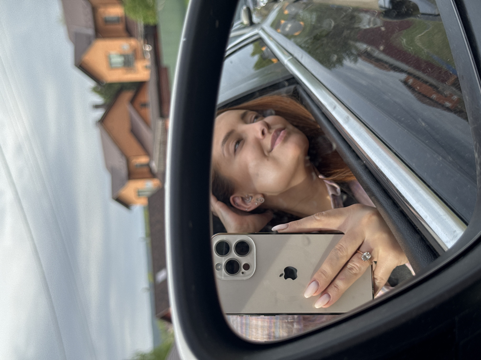 The image shows a woman reflected in a car's side mirror. She's holding a silver iPhone, capturing her own reflection. She has fair skin, reddish-brown hair, and is wearing a ring on her ring finger. The background visible through the mirror includes the interior of the car and an out-of-focus view of a house with a red roof. The sky appears cloudy. There is not enough information in the picture to indicate that the location is in Гавриловка, Russia.