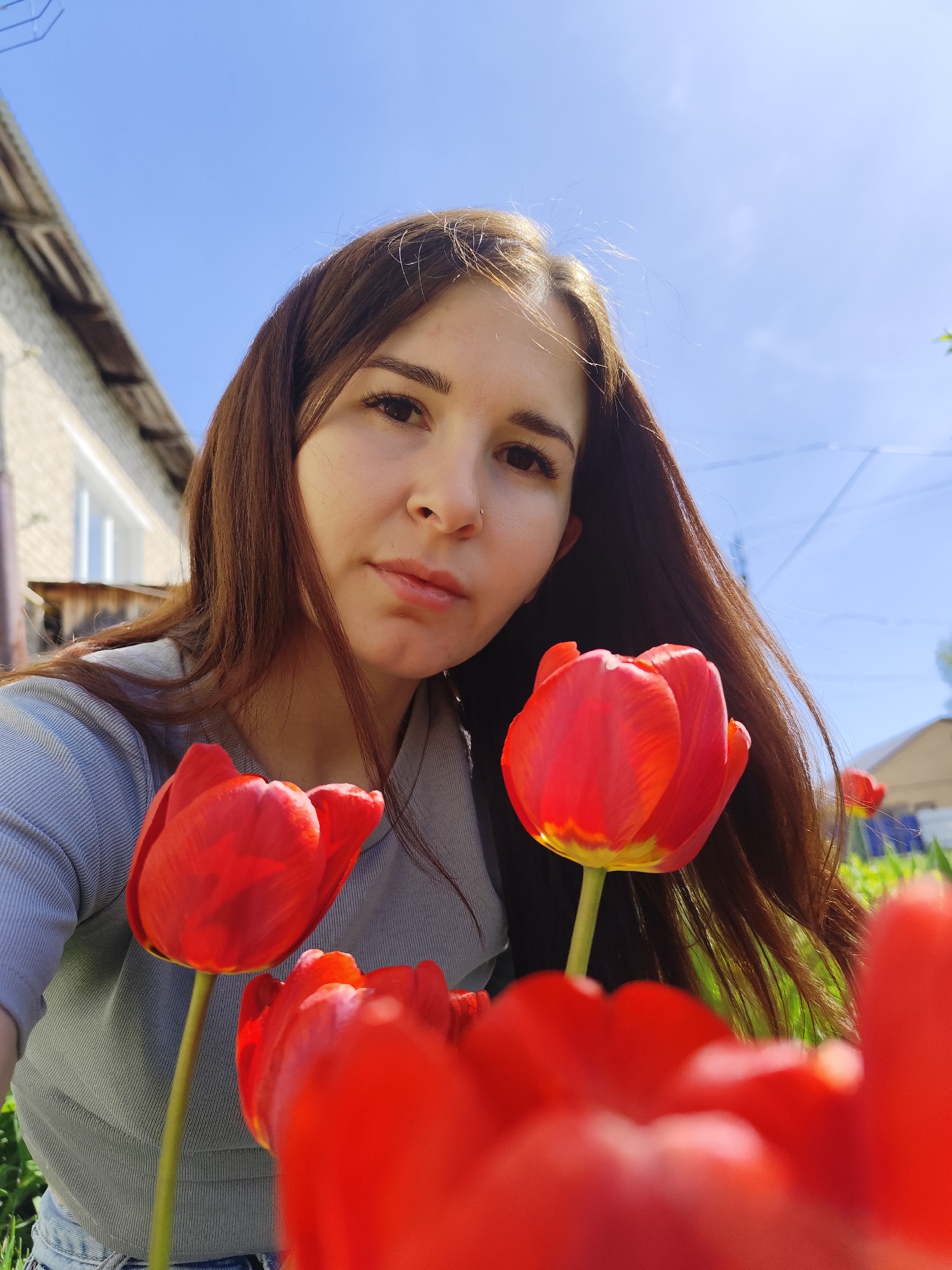 A young woman with long brown hair is centered in the frame, taking a selfie. She has fair skin, brown eyes, and a piercing in her nostril. She is wearing a gray shirt. In the foreground, several red tulips are prominently featured, some in focus and others slightly blurred, giving a shallow depth of field. Behind the woman, the blurred background suggests a residential area, with a partially visible house to the left and a clear blue sky overhead. The location is most likely in the city of Фоки, Russia.