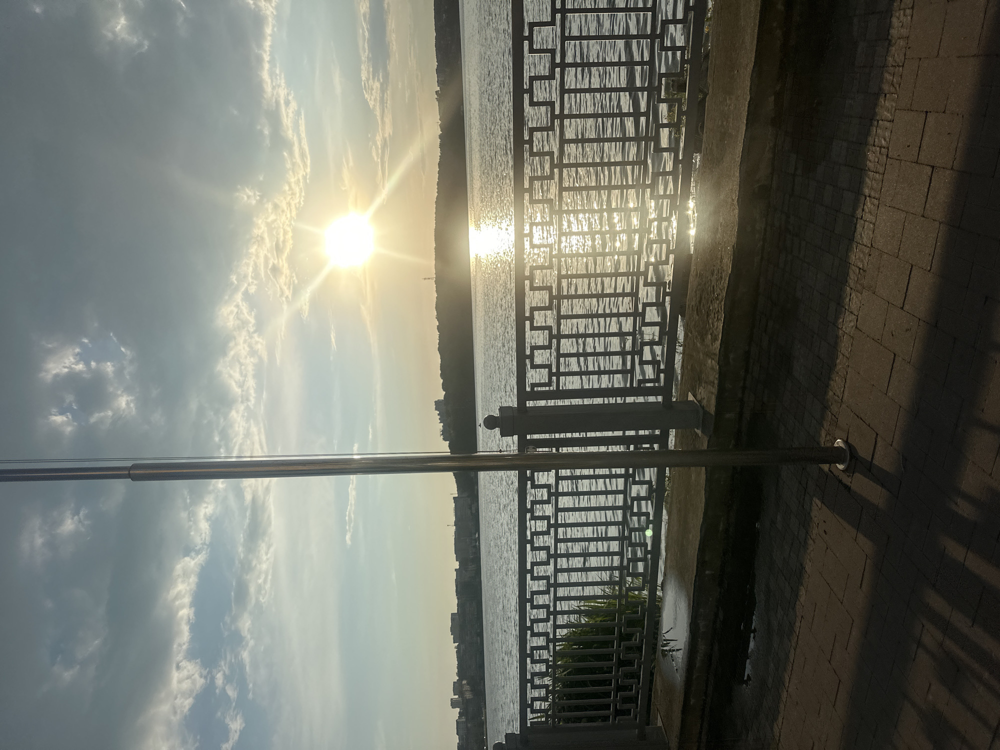This media file captures a scenic view of a waterfront promenade under a bright, sunny sky. The sun, positioned high in the sky, casts a brilliant glare across the water, creating a shimmering effect. Wispy clouds dot the sky, illuminated by the sun's rays.
The foreground features a decorative metal railing, its geometric pattern casting shadows on the paved walkway behind it. The walkway is made of brick or stone pavers. A large metal pole stands vertically, a prominent object in the scene.
Beyond the railing, a wide body of water stretches out, its surface rippled by the wind. In the distance, the silhouette of a city or town is visible along the opposite shore, with buildings lining the horizon. The overall scene suggests a pleasant day, likely in the late afternoon given the angle of the sun and the presence of shadows. The mood is serene and perhaps a little tranquil, with the natural beauty of the water and sky dominating the view. There are no visible people or text in the image. The location is identified as Ternopil, Ukraine.