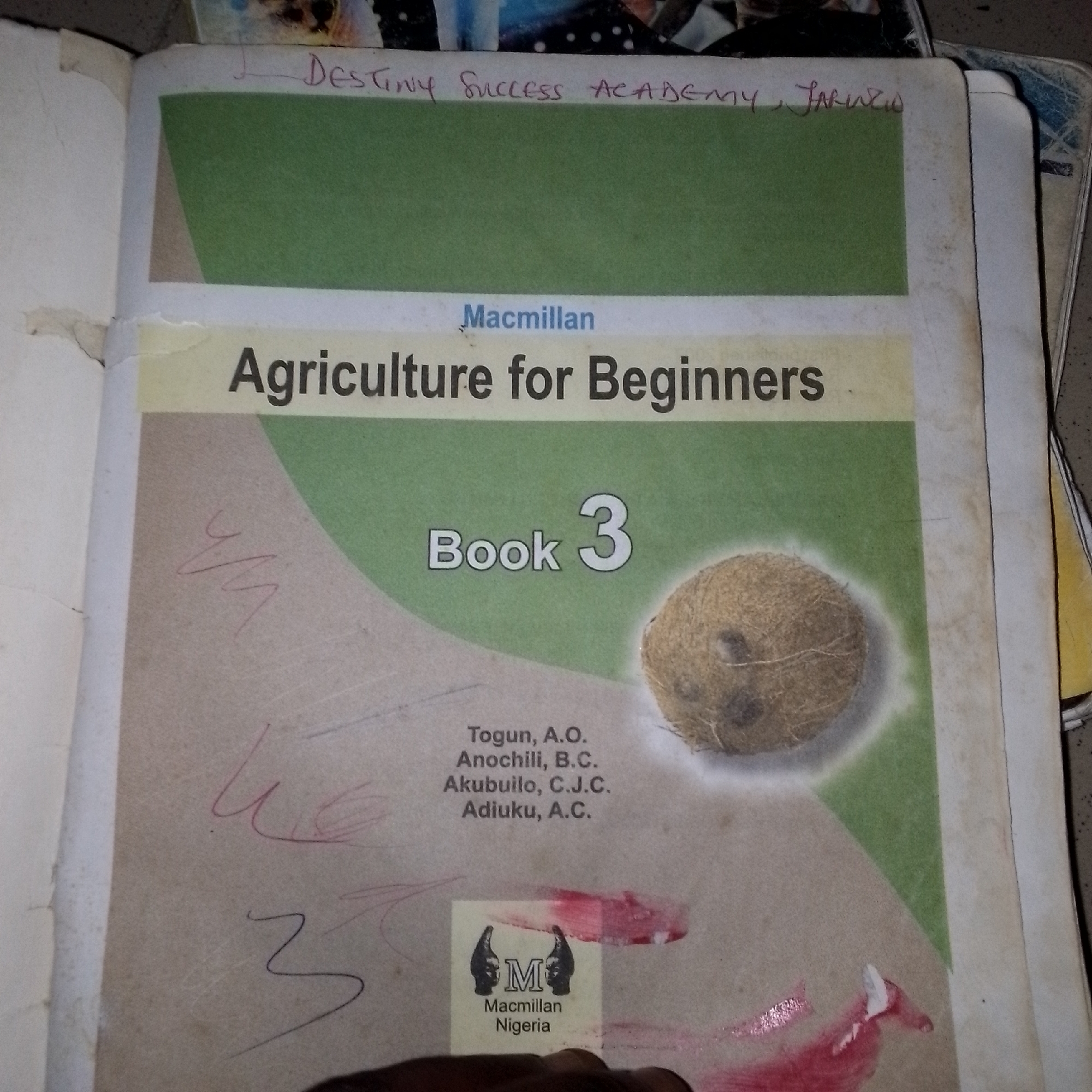 This is a close-up, eye-level photograph of an open book titled "Agriculture for Beginners Book 3" by Macmillan Nigeria. The book's cover features a green field and a picture of a coconut. Several names of authors are listed below the title: Togun, A.O.; Anochili, B.C.; Akubuilo, C.J.C.; and Adiuku, A.C. There are also handwritten annotations on the page, including "DESTINY SUCCESS ACADEMY, JALUNGO" in red ink at the top. Below the book, a person's hand is visible, obscuring part of the bottom of the image. The background shows other books and materials, suggesting a personal study or collection. The lighting is consistent, implying an indoor setting, likely during the day. The overall scene conveys a sense of learning and personal engagement with educational materials, with the handwritten notes adding a personal touch and indicating a connection to a specific academy in Jalingo, Nigeria.
