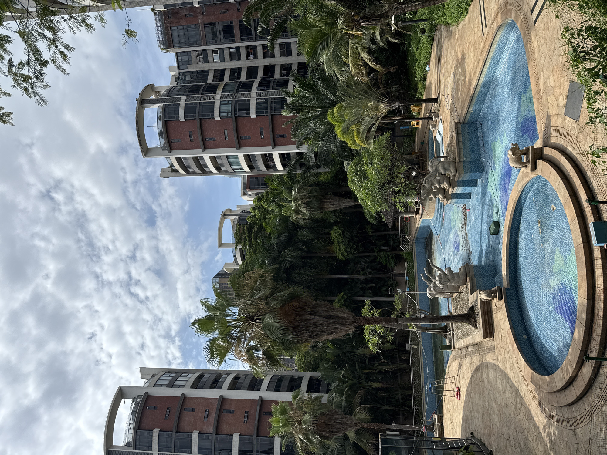 In Haizhu District, China, a resort-style swimming pool area is depicted, nestled between imposing modern apartment buildings. The pool itself is multi-leveled and partially filled with water, featuring intricate blue mosaic tiling that shifts in color. Sculptural elements, including what appear to be elephants and figures of people, are strategically placed around the pool's edges and on raised platforms. Surrounding the pool, paved pathways made of light-colored stone wind through lush, tropical vegetation, including numerous palm trees of varying sizes and other dense foliage. In the background, the architecture of the apartment buildings is characterized by brick facades, dark-paneled windows, and curved balconies, contrasted by white structural elements and circular top sections. The sky above is partly cloudy, with a mix of white and gray clouds against a blue backdrop. The scene appears quiet and deserted, with no individuals visible.