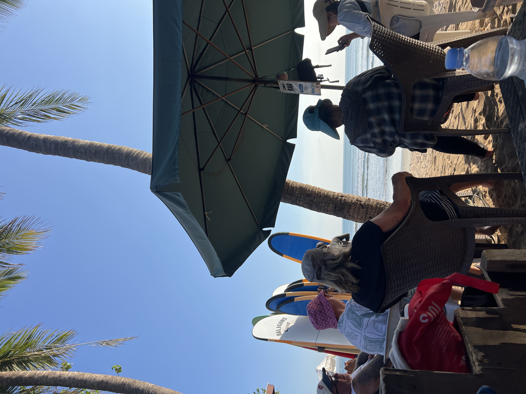 In Kuta, Indonesia, a group of people are seated on a sandy beach under a large green umbrella, facing the ocean. Several surfboards are propped up behind them, one with "BALIGUNS SURF" visible. To the right, a sign for a bar hangs from the umbrella. A woman on the far right is using her phone. In the foreground, a partially visible bottle of water rests on a wooden surface. Palm trees frame the scene against a clear blue sky.