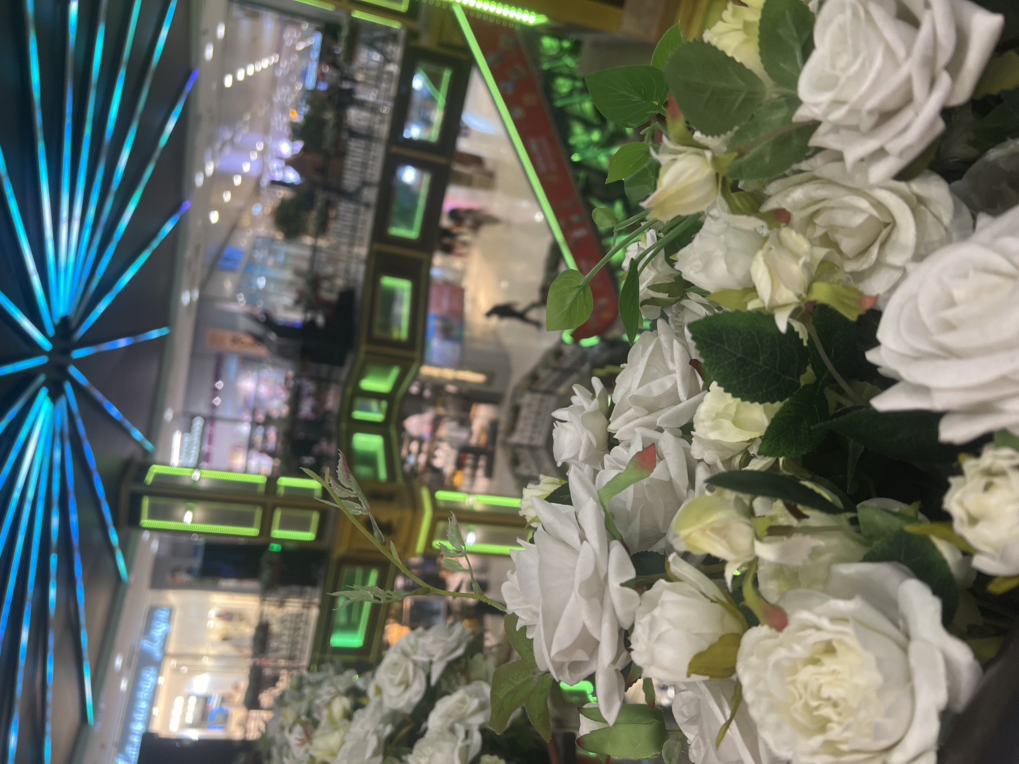 The image captures a scene with white roses in the foreground, possibly artificial, with green leaves interspersed. Behind the roses, a blurry reflection reveals an interior space with a Ferris wheel-like structure featuring blue lights. The architectural style and lighting suggest it could be a mall or entertainment complex. The store fronts are dimly lit. The reflection in the image also hints at the presence of people. Given the inquiry's context, it is implied that the location is potentially within a shopping center in Moscow, Russia.