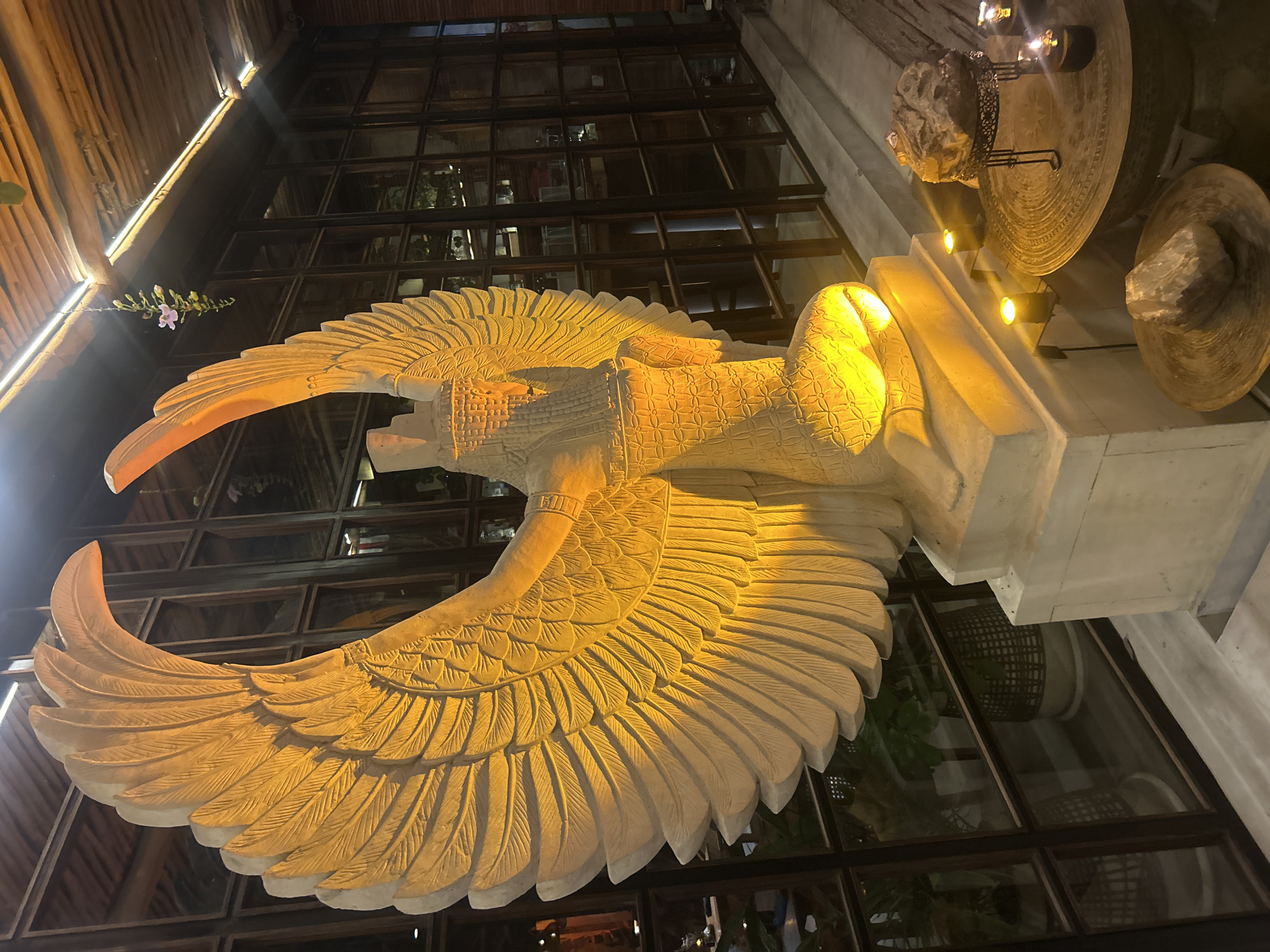 The image shows a large, light-colored statue of a winged humanoid figure kneeling. The statue is illuminated by warm yellow lights, highlighting the intricate carvings of feathers on its wings and the patterned clothing on its torso. The background features a large window with dark wooden frames, behind which are glimpses of potted plants. The floor in front of the statue has several small lit candles and decorative rocks on tables. The ceiling above is made of wooden beams with strip lighting. This statue is located in a restaurant in Mengwi, Indonesia.