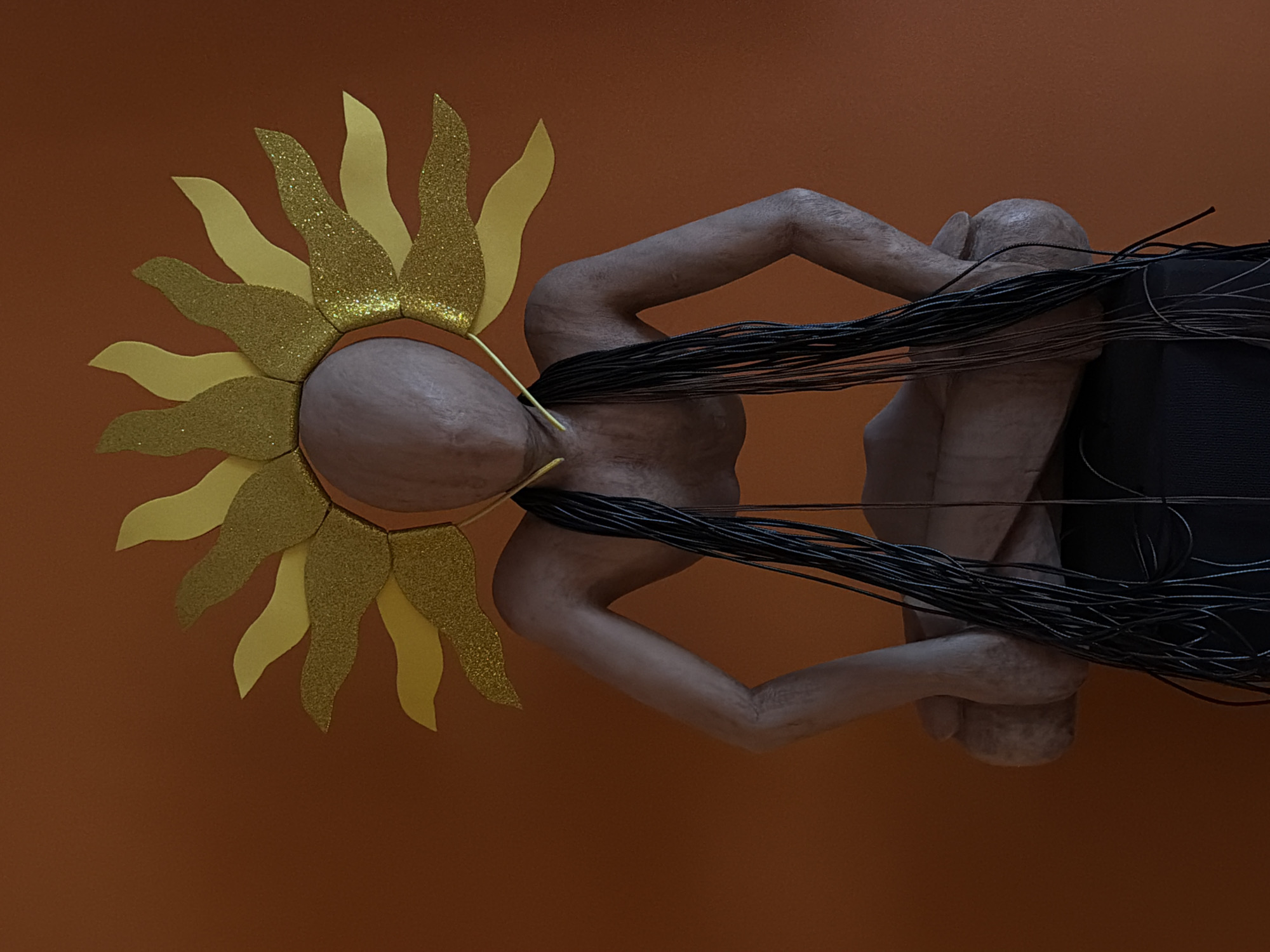 This is a digitally rendered image with an orange background. The central subject is a humanoid figure with tan skin, appearing to be in a fetal position. The figure has dark, string-like hair that emanates from its head and is draped over its body, extending to the right side of the frame. A golden sun-like headdress adorns the figure's head, with alternating matte gold and glittery gold sun rays. The figure's pose and the headdress create a stylized, almost ethereal representation. The overall mood is abstract and artistic, with no specific time of day, weather, or location cues discernible beyond the provided context. There is no visible text in the image.