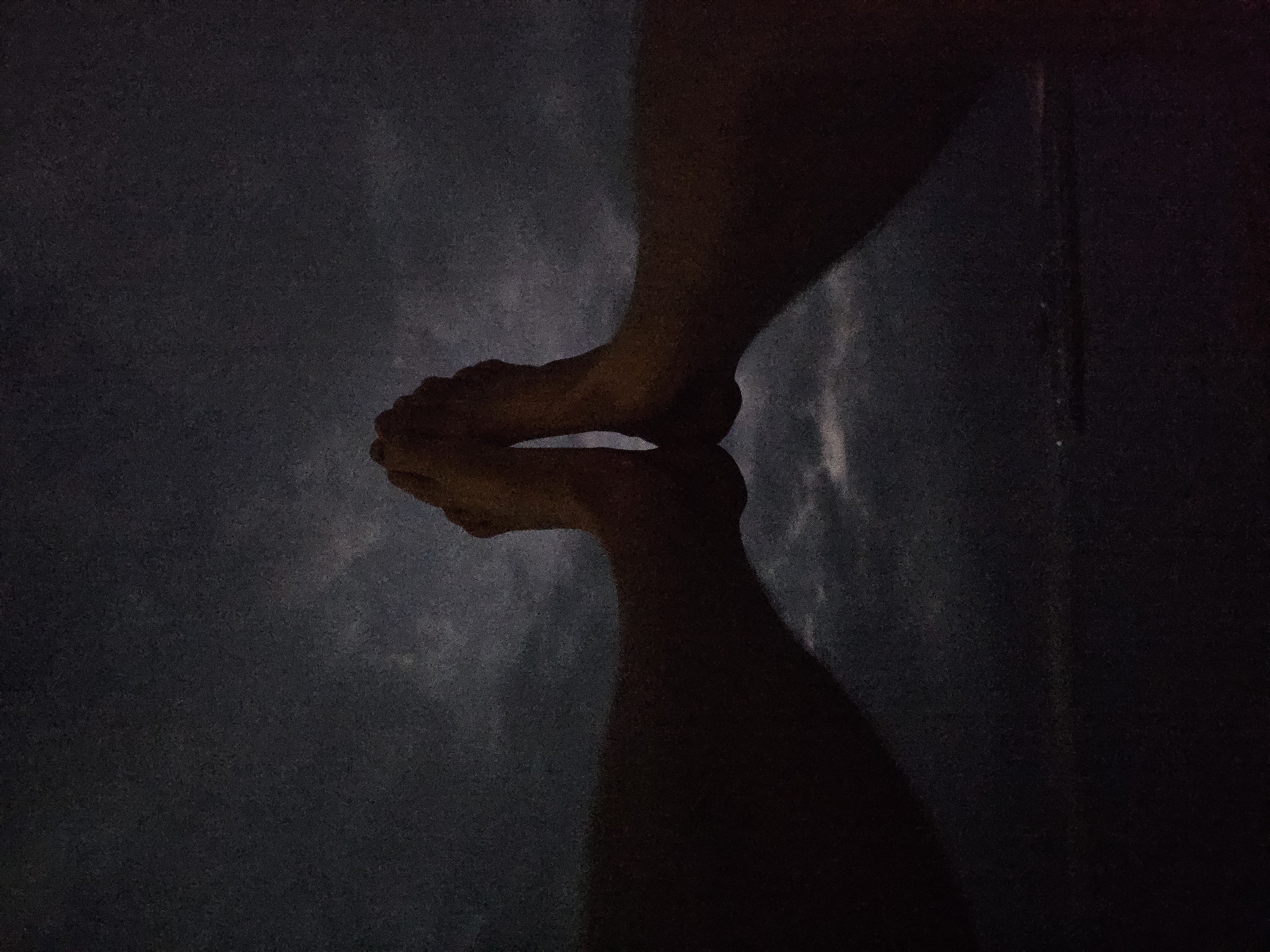 A dim, overhead shot shows two legs and feet, positioned in a way that creates a mirrored reflection on a surface. The lighting is poor, casting the scene in mostly dark tones. The surface upon which the legs stand appears to be water, possibly a swimming pool or similarly reflective body of water. The legs are light-skinned, and the reflection of the legs creates an almost symmetrical image. It is not possible to determine the location in Denpasar, Indonesia from this image.