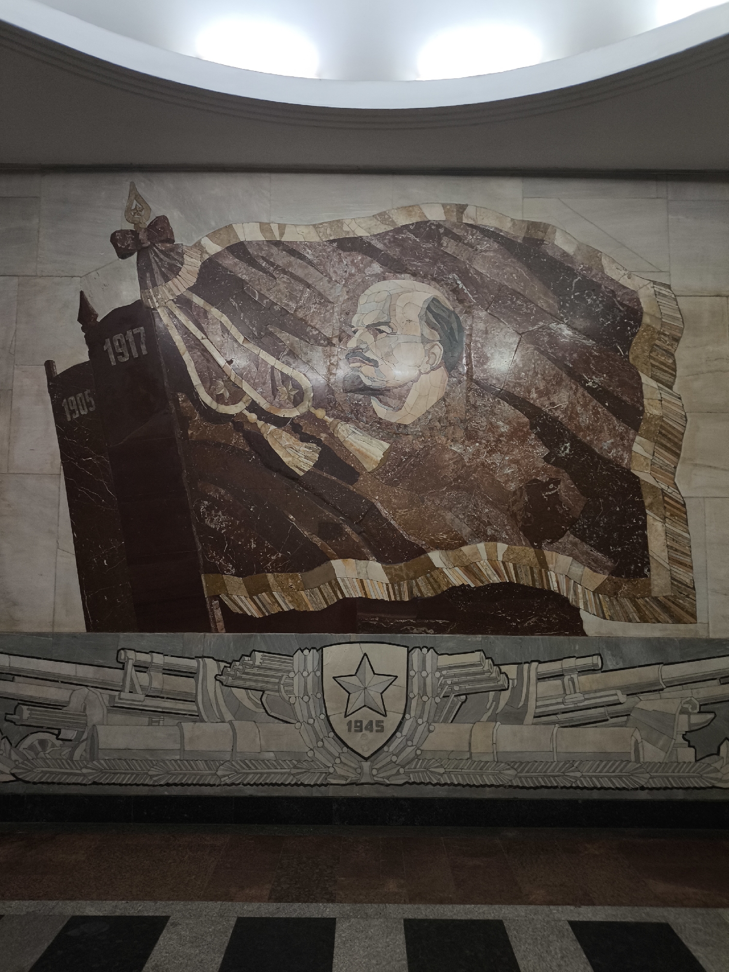 The image shows a mosaic mural on a wall. The mural depicts a flag with the dates 1905 and 1917 on it, with a profile portrait of Vladimir Lenin superimposed over the flag. Below the flag is a design incorporating cannons and a shield with a star and the date 1945. The location is not specified.