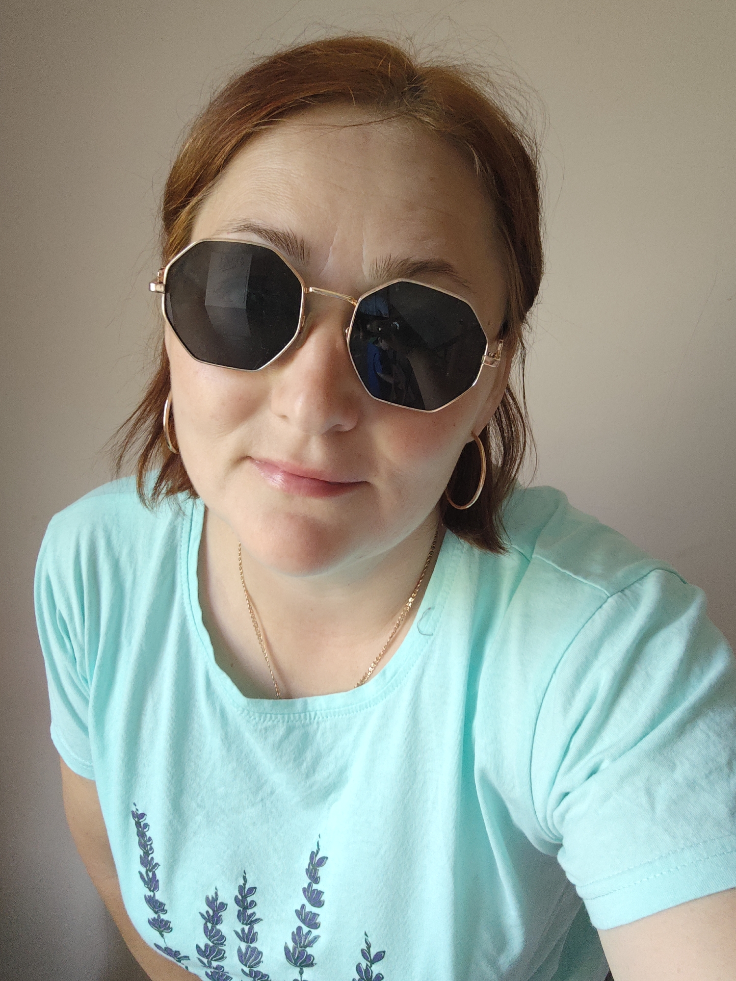A middle-aged woman with reddish-brown hair is posing for a selfie indoors. She is wearing large, gold-framed, dark sunglasses that obscure her eyes. She has a gold chain necklace and gold hoop earrings. She is wearing a light turquoise t-shirt with a floral design printed on the front. The background is a plain, light beige wall. The picture was not taken in Markovskii, Russia.