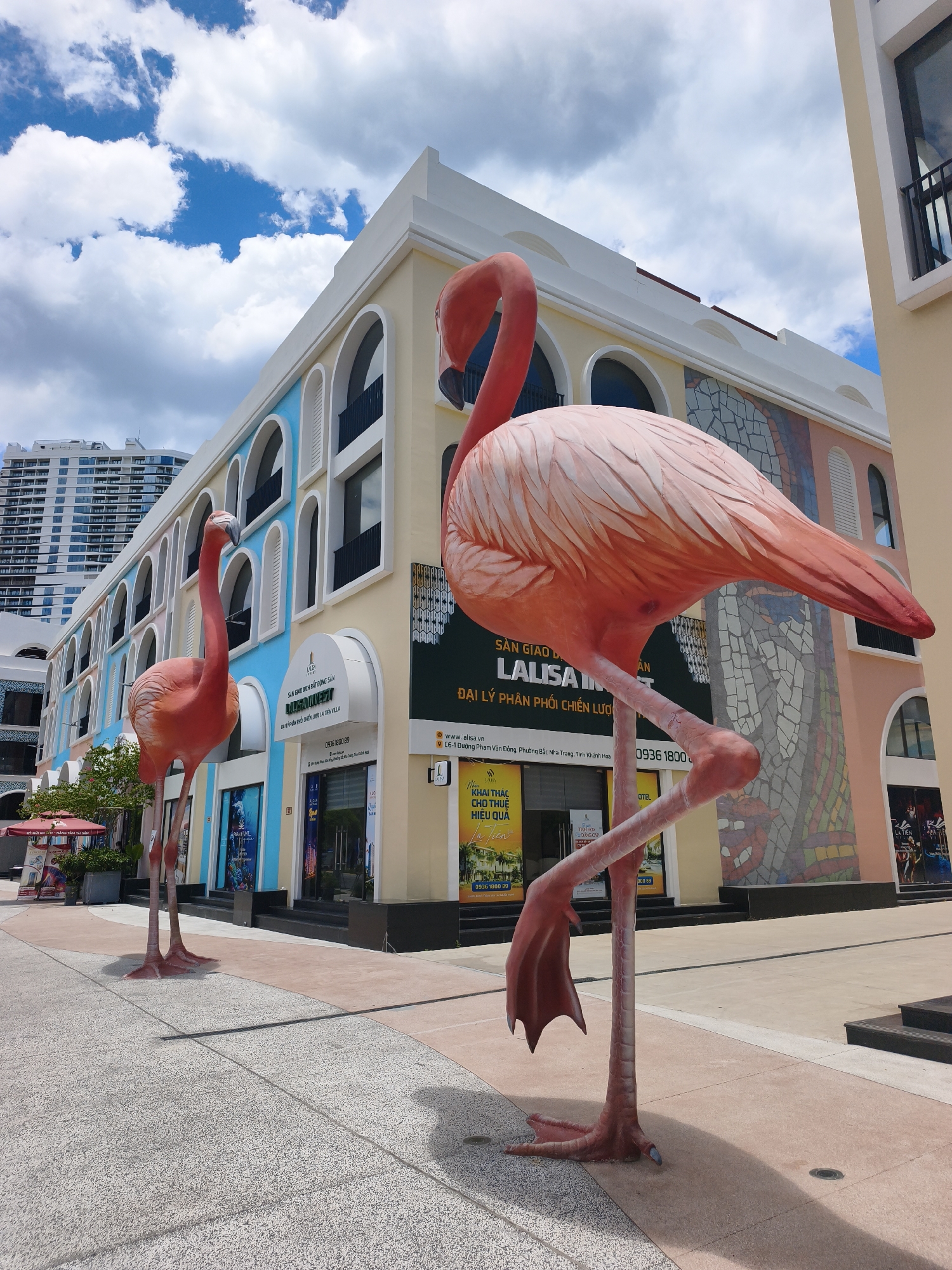 Two large, pink flamingo statues are positioned in the foreground of a modern building complex in Nha Trang, Vietnam. The building features a colorful facade with arched windows and sections of mosaic tile art. The flamingo on the right is closer to the camera and is in a standing pose with one leg raised. The flamingo on the left is smaller and further back, also standing. The ground in front of the building is paved with light-colored tiles and stone. In the background, a tall, multi-story building and a blue sky with white clouds are visible. Signs on the building indicate it is a commercial area, with one sign reading "LA LISSA INVESTMENT" and another advertising a "HOTEL." The overall scene suggests a public space, possibly a promenade or plaza in a resort or shopping area.