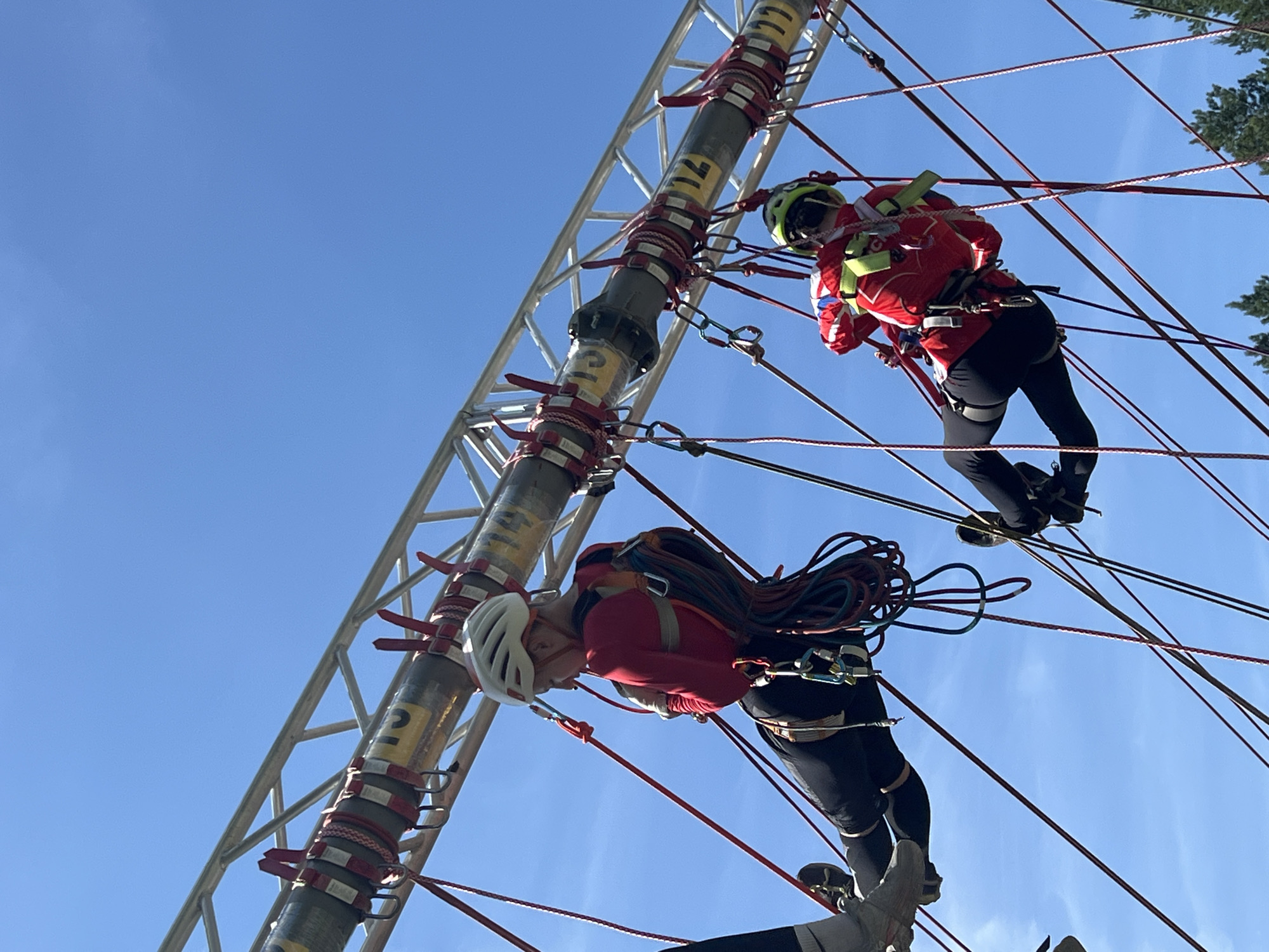 This is a low-angle, medium shot of two individuals suspended by ropes from a metal truss structure against a clear blue sky. Both individuals are wearing helmets and bright red jackets, suggesting they are participating in a recreational activity or training exercise. The person on the left, closer to the camera, is wearing a white helmet and appears to be looking down. The person on the right, further away, wears a lime green helmet and is also suspended by ropes. The truss structure is industrial in appearance with yellow markings on a pipe that is part of the setup. There are numerous red and other colored ropes crisscrossing the frame, indicating a complex rigging system. The background is a cloudless sky, with the tops of some evergreen trees visible at the bottom right of the image. There is no indication of the location being in the city of Penza, Russia.