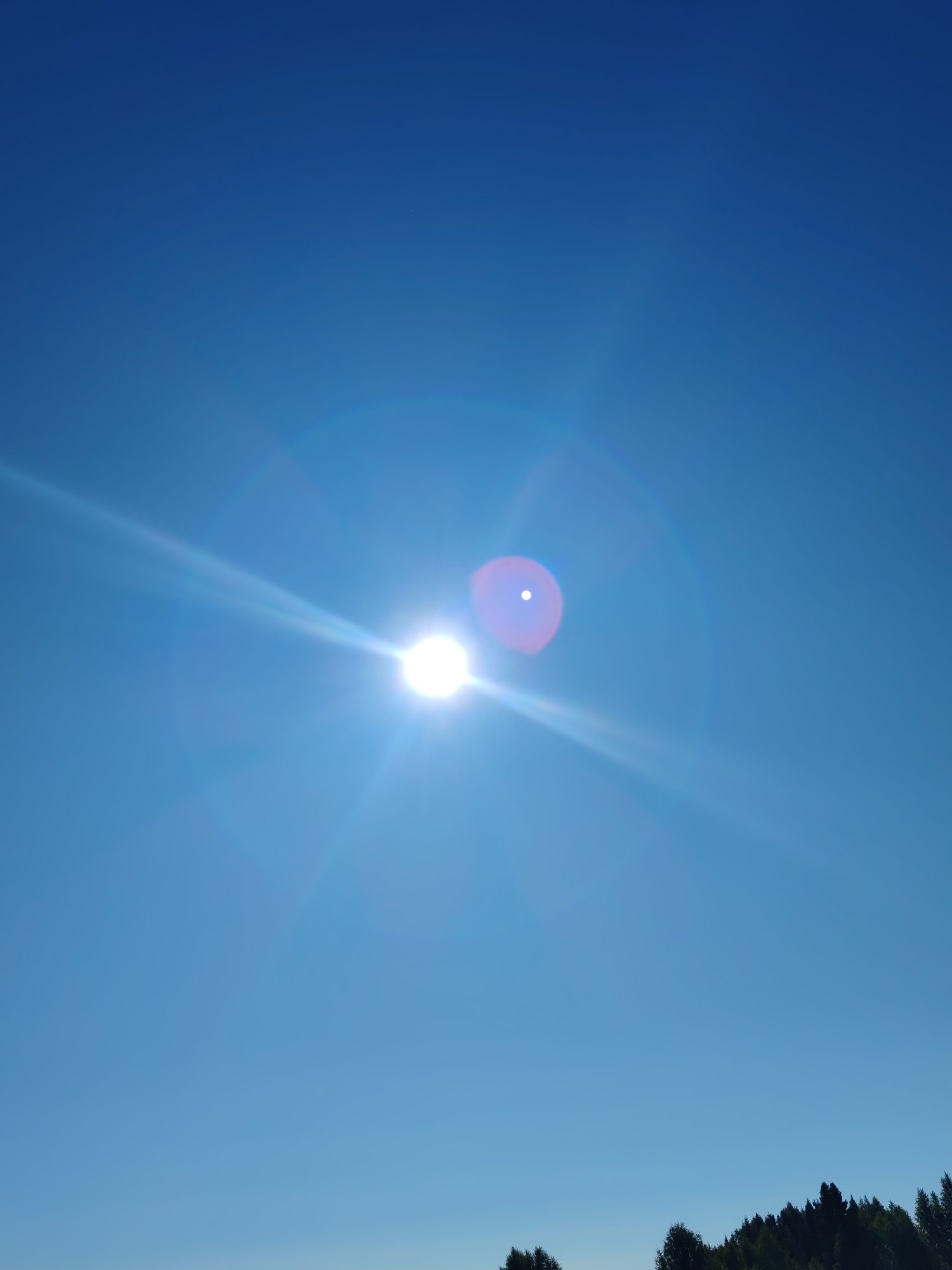 The image captures a bright, clear blue sky with the sun prominently featured. The sun appears as a radiant sphere with lens flare extending outwards in sharp rays. A distinct circular flare with a white dot in the center is visible to the left of the sun. The lower portion of the image shows the dark silhouette of a dense evergreen forest against the bright sky, suggesting a natural landscape. No people or specific urban elements are visible. The scene is likely an outdoor shot taken during daylight hours. It is not possible to determine the precise location within Чухлэм, Russia from the provided image.