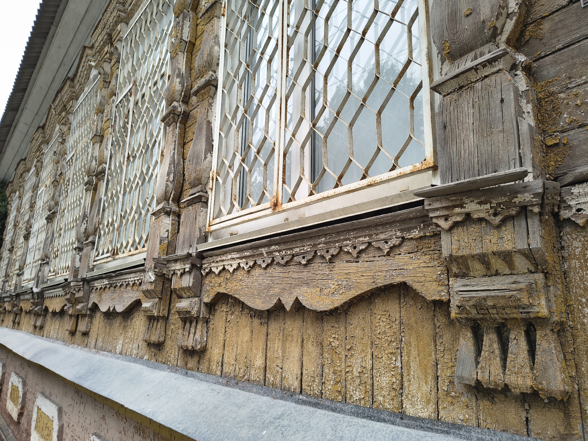 The image shows the facade of an old wooden building with ornate carved elements and barred windows. The wood is weathered and the yellow paint is peeling significantly, revealing the natural grain and age of the material. The windows have a diamond-shaped metal grating, suggesting a historical or functional purpose for security. The building appears to be a residential or public structure with multiple windows visible along its length. The architectural style suggests it could be from the late 19th or early 20th century, common in older towns in Russia. The lower part of the image shows a part of a stone foundation or wall with a decorative pattern, and a metal roof edge of an adjacent structure. The overall impression is one of historical preservation, with the building showing signs of age and possibly neglect. The location is specified as Mikhaylovka, Russia, a city known for its historical architecture. There are no people or animals visible in the image.