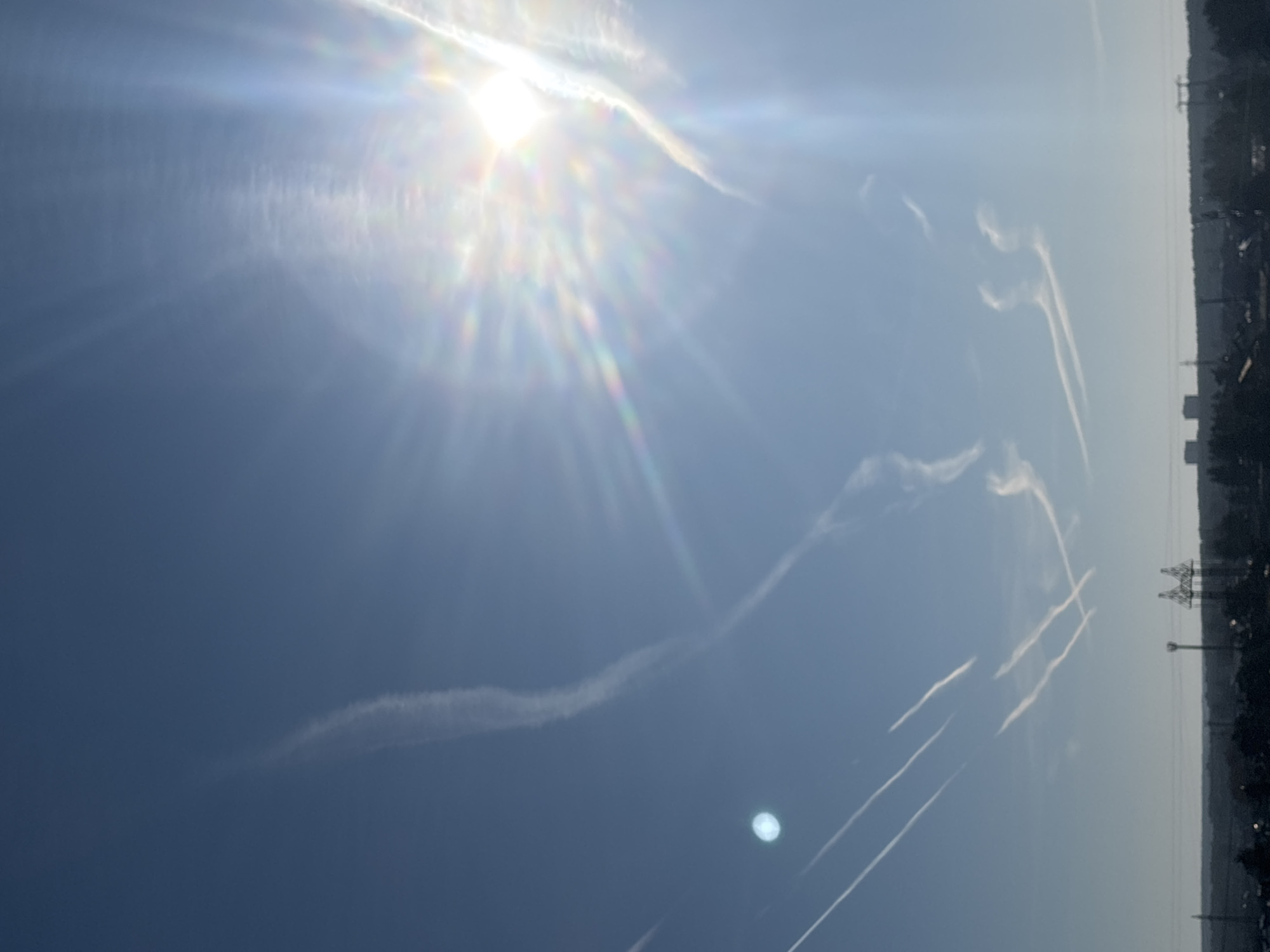 The image shows a sky with contrails, possibly above the city of Chaikovsky, Russia. The sun is bright and prominent, surrounded by radiating light and atmospheric effects. There are several distinct contrails visible against the blue sky, some linear and others curved, exhibiting signs of dispersion. The horizon line suggests a land mass with trees and what appears to be power lines and buildings.