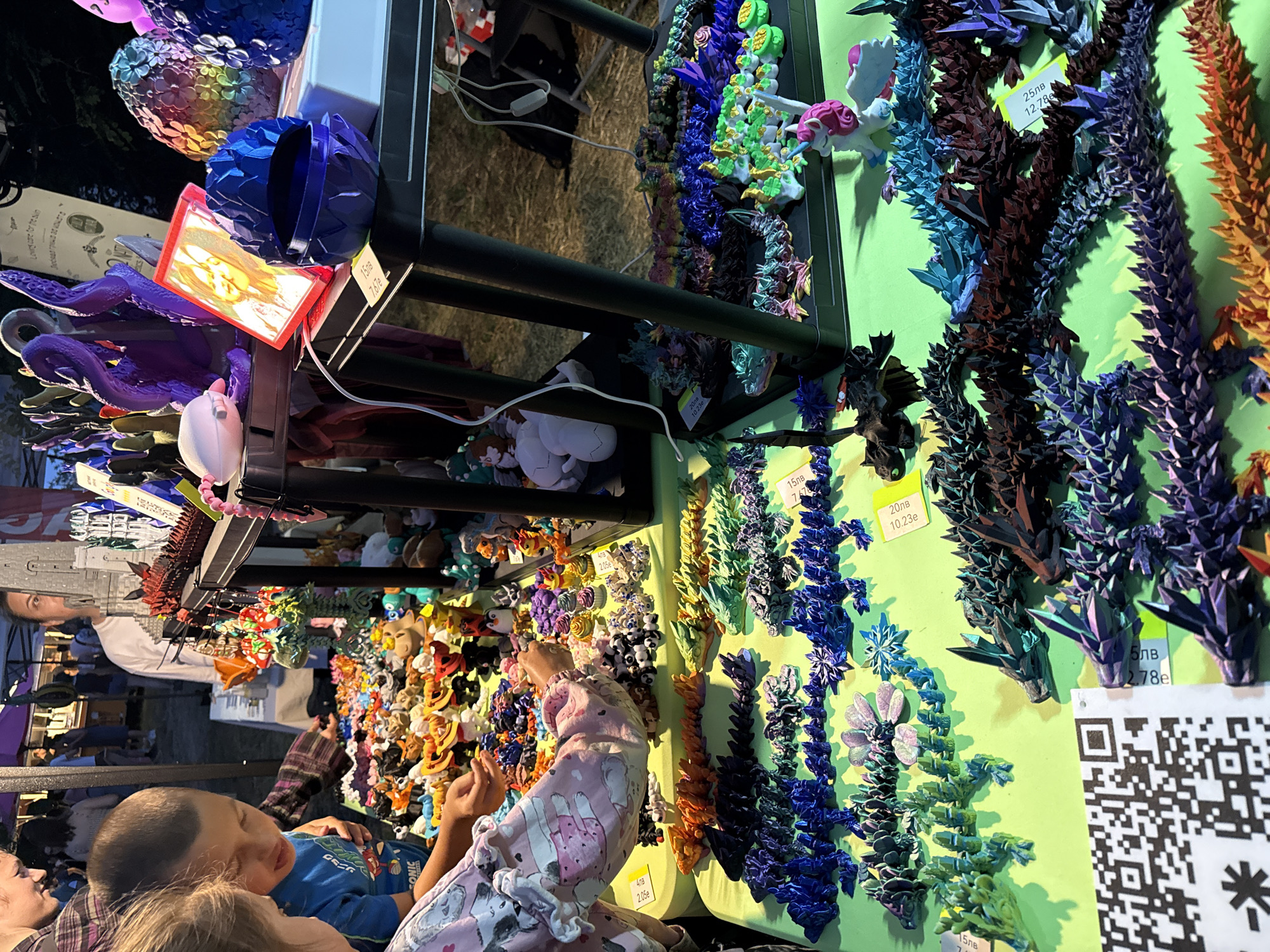 A photograph captures a bustling outdoor market scene, likely in Stara Zagora, Bulgaria, where various items are displayed for sale. In the foreground, a bright green table is laden with a multitude of colorful, intricately designed figurines, predominantly dragons and mythical creatures, arranged in rows. Some of these items are accompanied by small yellow price tags. A QR code is prominently placed on the left side of the table.
Two children are visible in the left portion of the frame, engaged with the displayed merchandise. A boy wearing a blue t-shirt with graphics is looking down, seemingly pointing at or examining some of the figurines. A young girl, wearing a pink pajama-like top with cartoon cat prints, is also looking at the items on the table, with her hand extended towards them.
In the background, several adults are present, some of whom are standing behind tables, possibly vendors. The atmosphere suggests a lively event, with tents or stalls set up. The lighting indicates it might be evening or dusk, with artificial lights illuminating the scene. Other objects on display include decorative eggs with intricate floral patterns, purple tentacle-like sculptures, and a framed picture. The overall scene depicts a vibrant marketplace with a focus on artisanal crafts or toys.