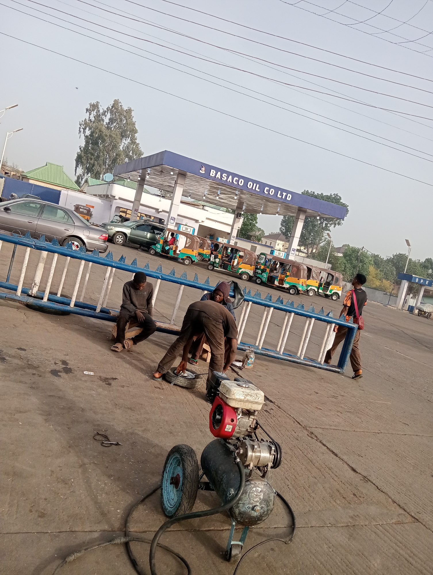 This image captures a bustling scene at a Basaco Oil Co Ltd fuel station in Jos, Nigeria. In the foreground, several men are engaged in activity near a blue and white metal barrier. One man, dressed in a dark sweatshirt and trousers, sits on a small wooden bench next to the barrier. Another man, wearing dark clothing and sandals, is bent over, actively working on a tire with a tool, suggesting a tire repair or inflation service. An air compressor, featuring a red and white engine on top of a black tank with a blue wheel, sits on the ground next to him, connected by hoses. A third man, carrying a bag, walks towards the right side of the frame, looking away from the camera.
In the background, behind the barrier, are several vehicles, including a silver sedan and a dark green SUV, as well as a line of colorful auto-rickshaws (commonly known as Keke Napep in Nigeria) parked near the fuel pumps. The gas station canopy prominently displays "BASACO OIL CO LTD". On the far right, a pump structure has "DPK" visible, likely indicating Dual Purpose Kerosene. The sky is bright, suggesting daytime, possibly late morning or early afternoon, with power lines stretching across the upper part of the frame. Trees and other buildings are visible further in the background. The scene depicts a typical everyday service stop in an urban Nigerian setting.