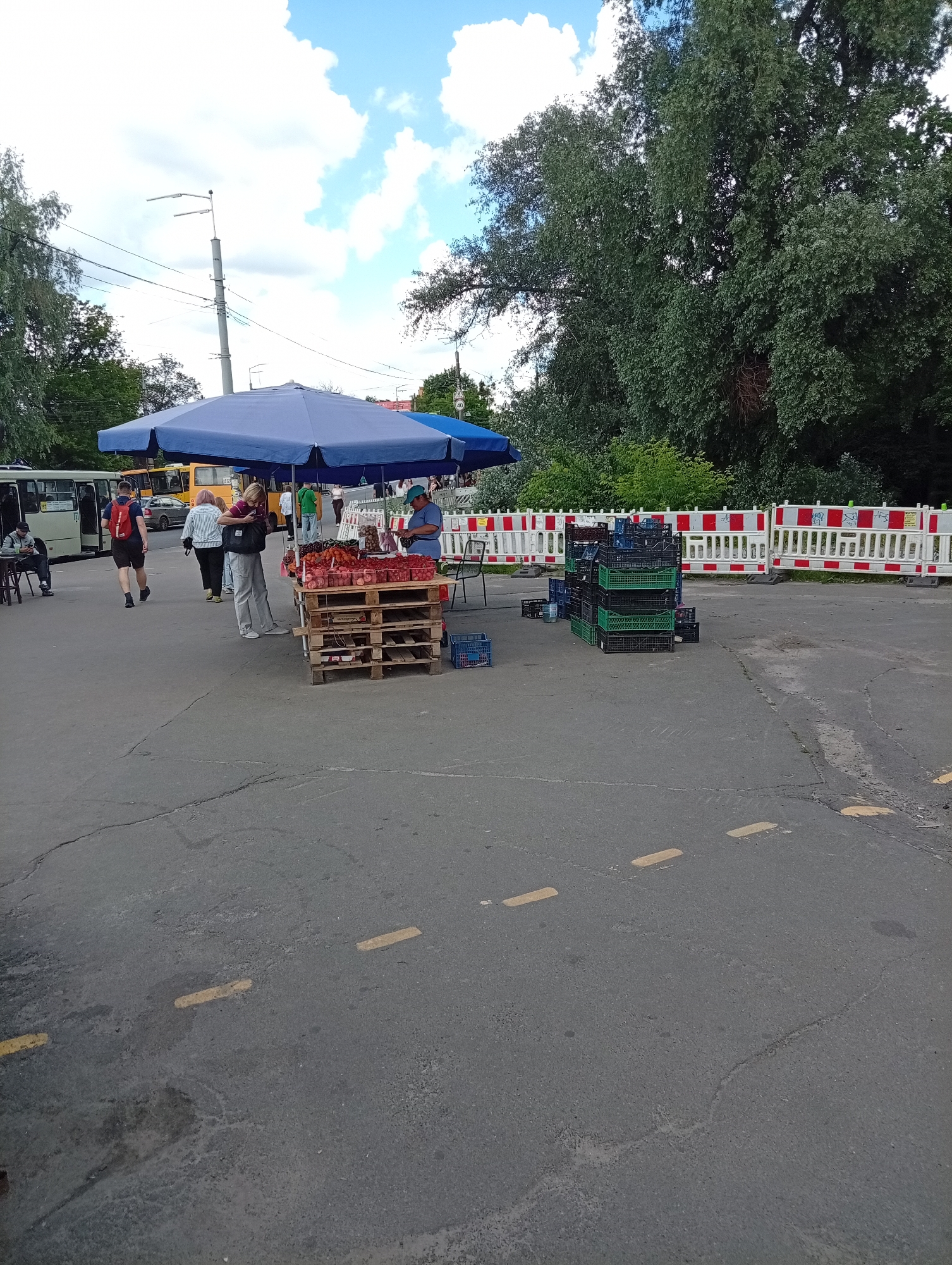 This media file depicts an outdoor market scene, possibly in a city or town, on a day with a partly cloudy sky. The foreground is dominated by a cracked asphalt surface marked with yellow lines.
Several people are present. On the left, a man with a red backpack walks away from the camera, and a woman in light-colored pants and a dark top stands near a market stall, looking at her phone. Further back, other individuals are visible, including a person sitting at a table and two more walking. A vendor, wearing a blue shirt and hat, is seen at the market stall, which displays an assortment of fruits, including tomatoes and other red produce, arranged on wooden pallets.
The market stall itself is shaded by a large blue umbrella. To the right of the stall, stacks of black and green plastic crates are piled on the ground. In the background, a white and red striped barrier, possibly indicating construction or a restricted area, stretches across the scene. Behind the barrier, lush green trees form a dense backdrop. Several buses are parked or driving in the background, suggesting a transportation hub or a busy street nearby. The time of day appears to be daytime, likely late morning or afternoon, given the lighting. The overall atmosphere seems casual and functional, typical of a local marketplace.