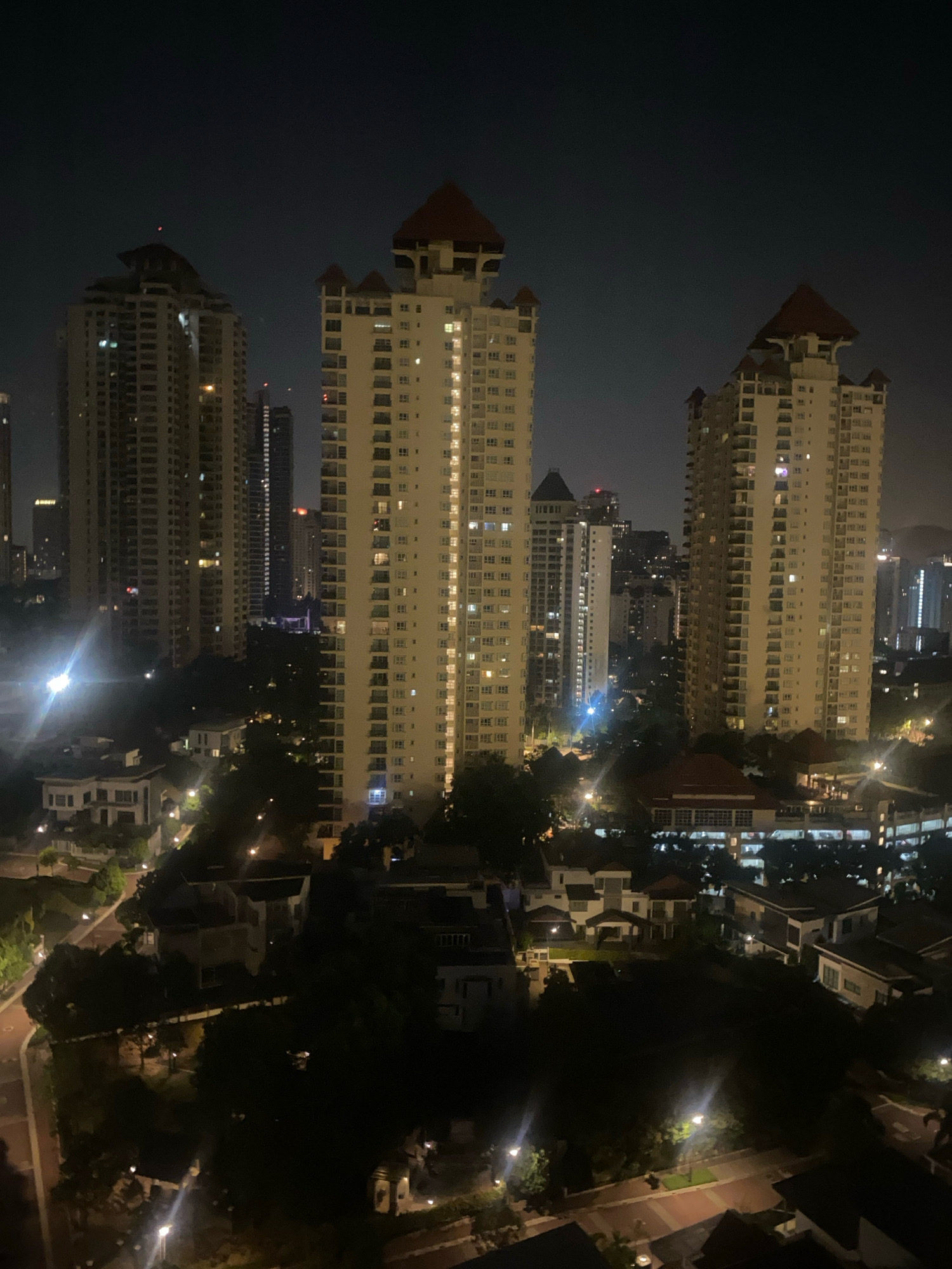 A nighttime, high-angle drone shot captures a cluster of residential high-rise buildings and lower-rise housing in Kuala Lumpur, Malaysia. The three prominent high-rises in the foreground are modern, beige-colored apartment buildings with pointed, red-tiled roofs. Each building features numerous illuminated windows, indicating occupied units. The surrounding area is densely populated with trees and smaller residential structures, also dotted with lights. In the distance, more city lights and taller skyscrapers are visible, suggesting a bustling urban environment. The sky is dark, characteristic of nighttime. There are no people or active interactions visible in the scene. The overall impression is of a well-lit, residential part of a large city at night.