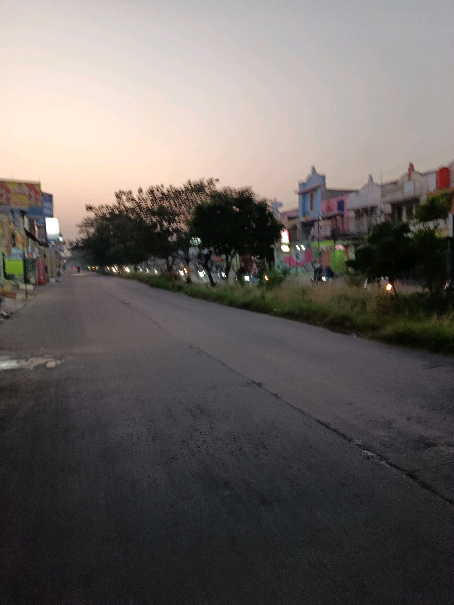 The image depicts a street scene in Bekasi, Indonesia, at dusk. The sky is a pale orange and pink hue, indicating the setting sun. The road, a dark asphalt surface, occupies the majority of the foreground. On the left side of the road, several buildings are visible, some adorned with colorful signage. The buildings appear to be shops or residences. Trees with sparse foliage line the right side of the road, with some streetlights or lamps illuminating the area. In the background, more buildings and trees are visible, shrouded in the dim light of dusk. A few indistinct figures or vehicles can be seen on the road, and the overall impression is one of a quiet, early evening in a city environment.