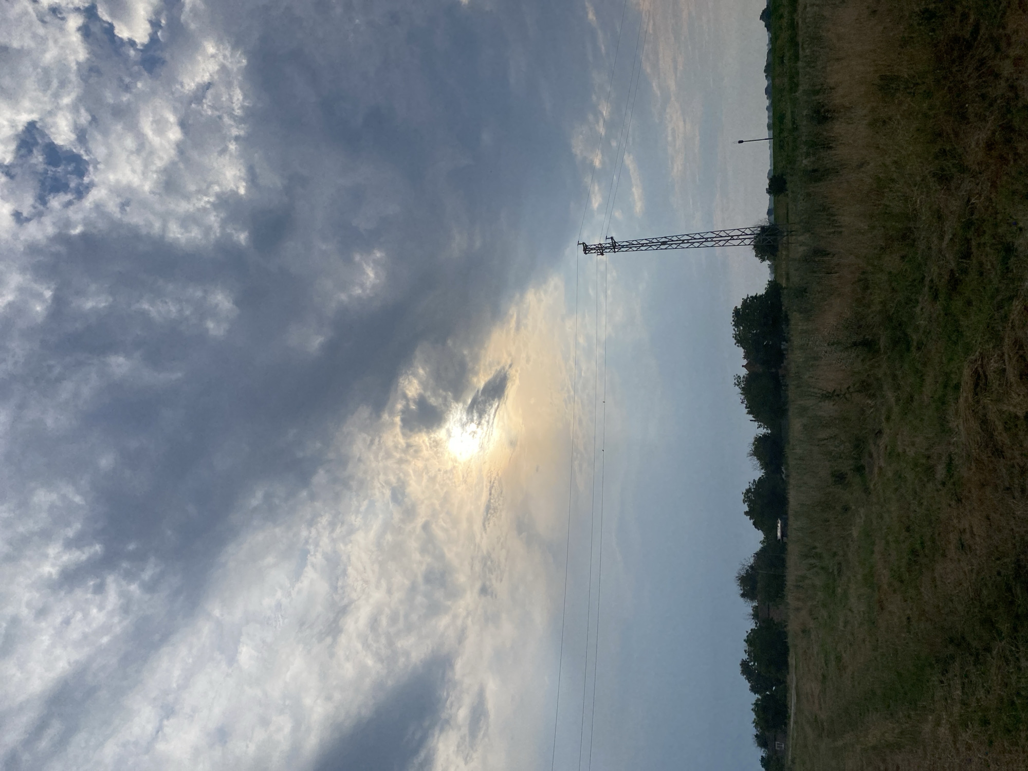 A wide, low-angle shot captures a field in Brashlen, Bulgaria, under a dramatic sky. The sun, partially obscured by clouds, shines through a break in the overcast sky, casting a warm glow on the upper portion of the frame. A tall, slender metal tower stands in the mid-ground, with three parallel power lines stretching horizontally across the sky in front of it. The foreground is dominated by dry, tall grass, some of which appears to have been recently cut. In the distance, a line of trees separates the field from what appears to be a small village or scattered buildings. The overall impression is one of rural landscape bathed in the soft light of a cloudy day.