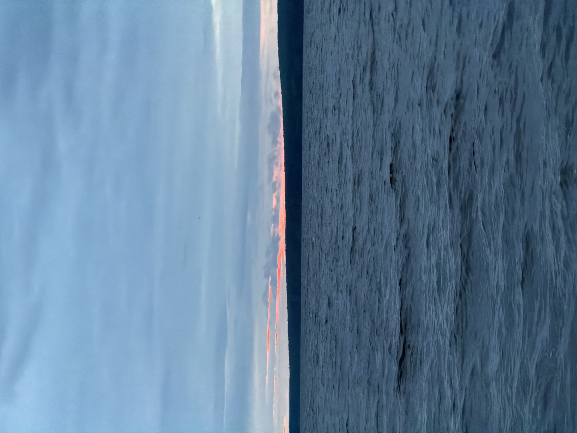This vertical image depicts a vast natural landscape dominated by a large body of water in the foreground and midground, under an overcast sky, observed in Chaikovsky, Russia. The lower two-thirds of the frame are occupied by dark blue-grey water, exhibiting a textured surface with numerous ripples and small waves, indicative of movement. A low, dark horizontal silhouette of a distant landmass, likely a forested shoreline or hills, spans the horizon line, separating the water from the sky. Immediately above this distant landmass, a narrow band of the sky displays distinct, elongated horizontal streaks of vibrant pink and orange light, suggesting either sunrise or sunset. The upper portion of the image, comprising the majority of the sky, is covered by a dense, uniform layer of grey-blue clouds with minimal visible structure. The overall lighting is dim and diffused. No individuals, vessels, or man-made objects are discernible within the frame.