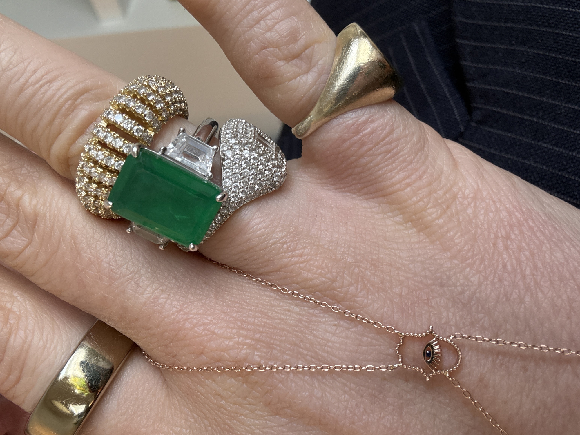 The image displays a close-up, top-down view of the back of a left human hand, adorned with multiple items of jewelry. The hand is fair-skinned, with visible skin texture.
On the index finger, a wide, plain gold-toned band with minor surface abrasions is worn. The middle finger wears three rings:
1. A wide, gold-toned band featuring multiple rows of pavé-set clear, round stones.
2. Below it, a silver-toned ring centered with a large rectangular emerald-cut green stone, flanked by two smaller rectangular clear stones.
3. Beneath that, a silver-toned ring with a domed, textured surface entirely pavé-set with numerous small clear, round stones.
The ring finger displays a distinct, irregularly shaped, polished gold-toned ring with a flared, organic form.
A delicate rose gold-toned chain, identified as part of a hand chain, runs across the back of the hand. It features a small Hamsa hand charm with a central evil eye motif, positioned between the index and middle fingers.
In the lower right background, partially visible beneath the ring finger, is a dark navy or black fabric with fine white pinstripes, indicative of tailored clothing. Reflections in the plain gold band on the index finger show indistinct light and dark shapes. No visual information within the image provides details sufficient to identify a specific location in the city of London, United Kingdom.