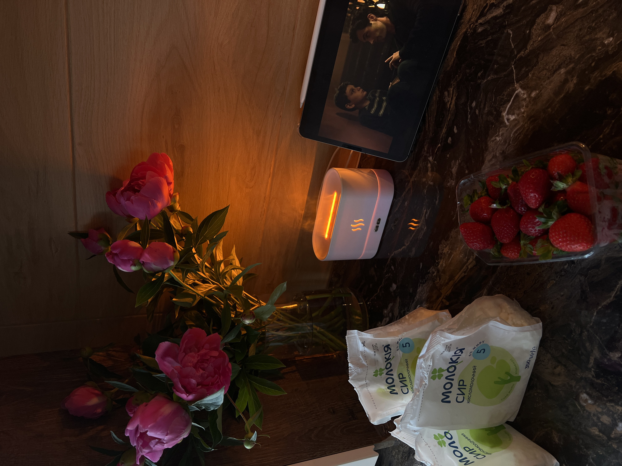The image depicts an indoor scene, likely a kitchen or dining area, situated in Ternopil, Ukraine. The primary elements are arranged on a dark, reflective, possibly marbled countertop beneath a light-colored wooden wall.
On the far left, a clear glass vase holds a bouquet of approximately ten vibrant pink peonies, some in full bloom and others as closed buds, with dark green foliage. Water is visible within the vase.
To the left-front, three white plastic packages are stacked, partially obscuring one another. Each package bears the Ukrainian brand name "МОЛОКІЯ" (Molokiya), identified as "СИР КИСЛОМОЛОЧНИЙ" (fermented milk curd cheese) with "5% ЖИРУ" (5% fat). One package clearly displays a production date of "18.06.2020" and a time "09:42:23". The packages are bulging with their contents, consistent with curd cheese. Molokiya is a known dairy brand originating from Ternopil, Ukraine.
In the middle-right of the countertop, a white, rectangular electronic device, possibly an aroma diffuser or humidifier, emits a prominent orange glow from its top opening and features three smaller, illuminated flame-like symbols on its front face.
To the right of the electronic device, a silver-backed tablet, positioned at an angle, displays a video. The screen shows two male individuals in an indoor setting: a younger boy on the left and an adult man on the right, both appearing to be engaged in conversation or interaction. A white stylus, consistent with an Apple Pencil, rests horizontally on the top bezel of the tablet.
In the bottom-right corner, a clear plastic punnet contains a collection of fresh, bright red strawberries, some with their green calyxes still attached.
The background consists of light-colored wooden paneling or a wooden surface, extending upwards from the countertop. The overall lighting is warm and dim, creating a cozy atmosphere, with light sources contributing an orange hue to the scene and reflections on the dark countertop. There are no individuals physically present in the immediate scene, but human figures are visible on the tablet screen.