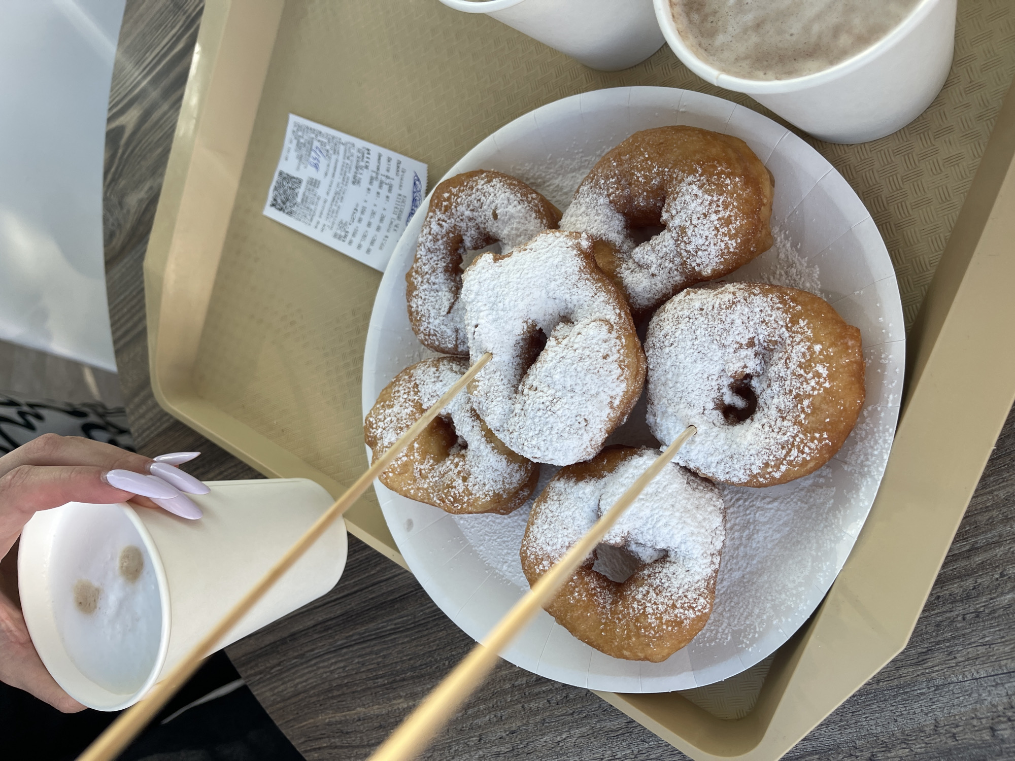 On a wooden table, a tan tray holds a white paper plate with six powdered doughnuts, a white paper cup of coffee, and a receipt. Two wooden skewers are stuck into the doughnuts. A person's hand with long, light purple nails holds the coffee cup, which is topped with frothed milk and two small brown dots. The receipt, crumpled slightly, displays Russian text and a QR code. In the background, a blurry white object, likely another cup, is visible. The scene is indoors, possibly in a cafe or diner, with the mention of Zelenogradsk, Russia, providing a specific location context.