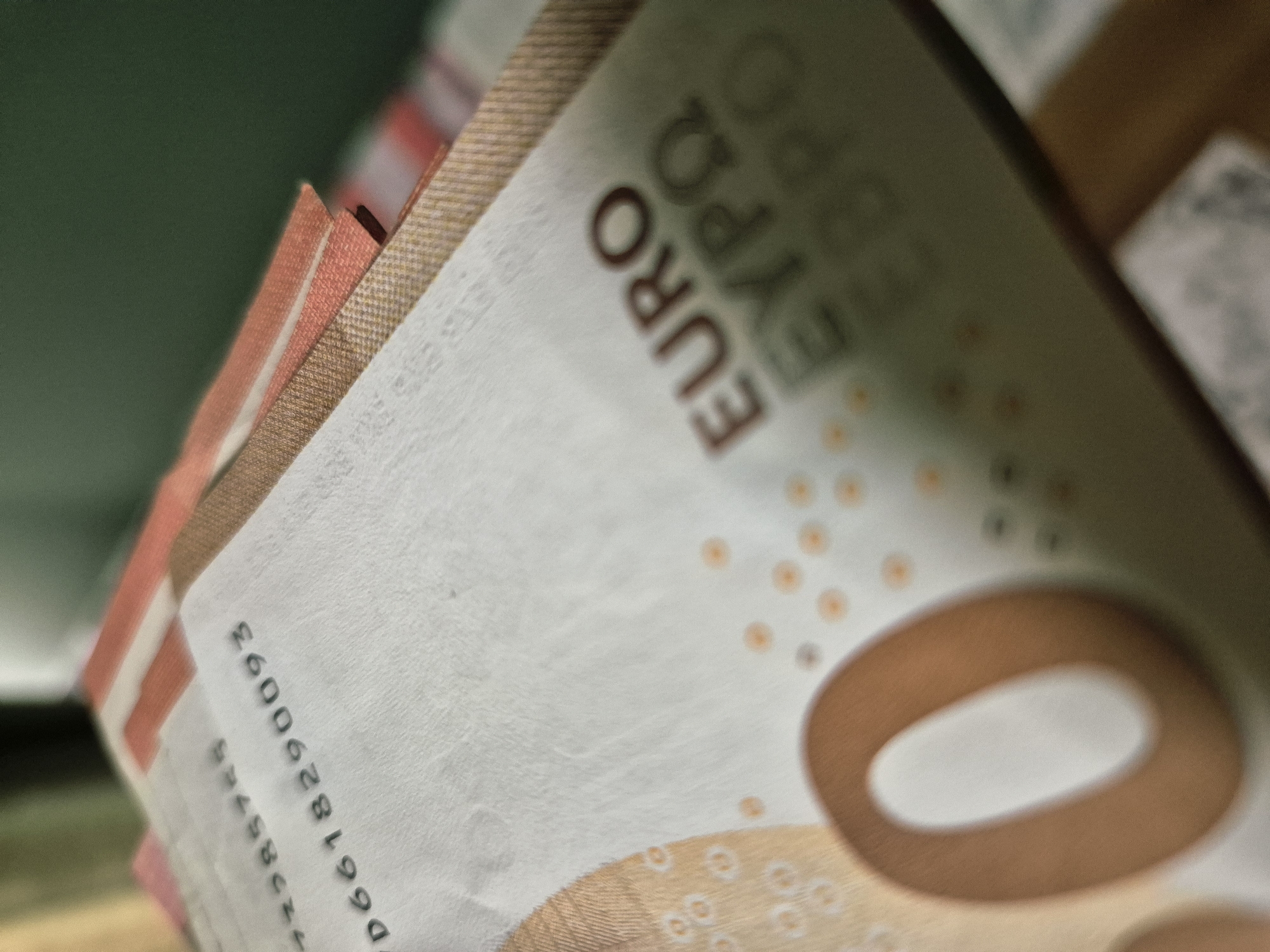 The scene depicts a close-up, low-angle view of a stack of Euro banknotes, situated indoors in Iskrets, Bulgaria. The uppermost banknote, identified as a 50 Euro note of the Europa series, is the primary subject. Its serial number, "D6618290093," is sharply focused and centrally located. A partial, out-of-focus number "2285785" is visible above it. The word "EURO" is clearly legible in dark brown text on the right side of the note, with the Greek characters "EYPΩ" and Cyrillic "EBPO" visible beneath it, slightly out of focus. A large, stylized numeral "0" (part of the "50" denomination) is prominent on the lower left portion of the note. Various security features and design elements, including small, circular dots, are visible on the banknote's surface. Below the top note, the textured, orange-brown edges of several other stacked Euro banknotes are partially visible, indicating a quantity of currency. The background is completely out of focus, presenting as indistinct fields of dark olive-green in the upper section and a lighter tan or brown color in the lower right, consistent with an indoor environment. No individuals, specific actions, or interactions are visible within the frame.