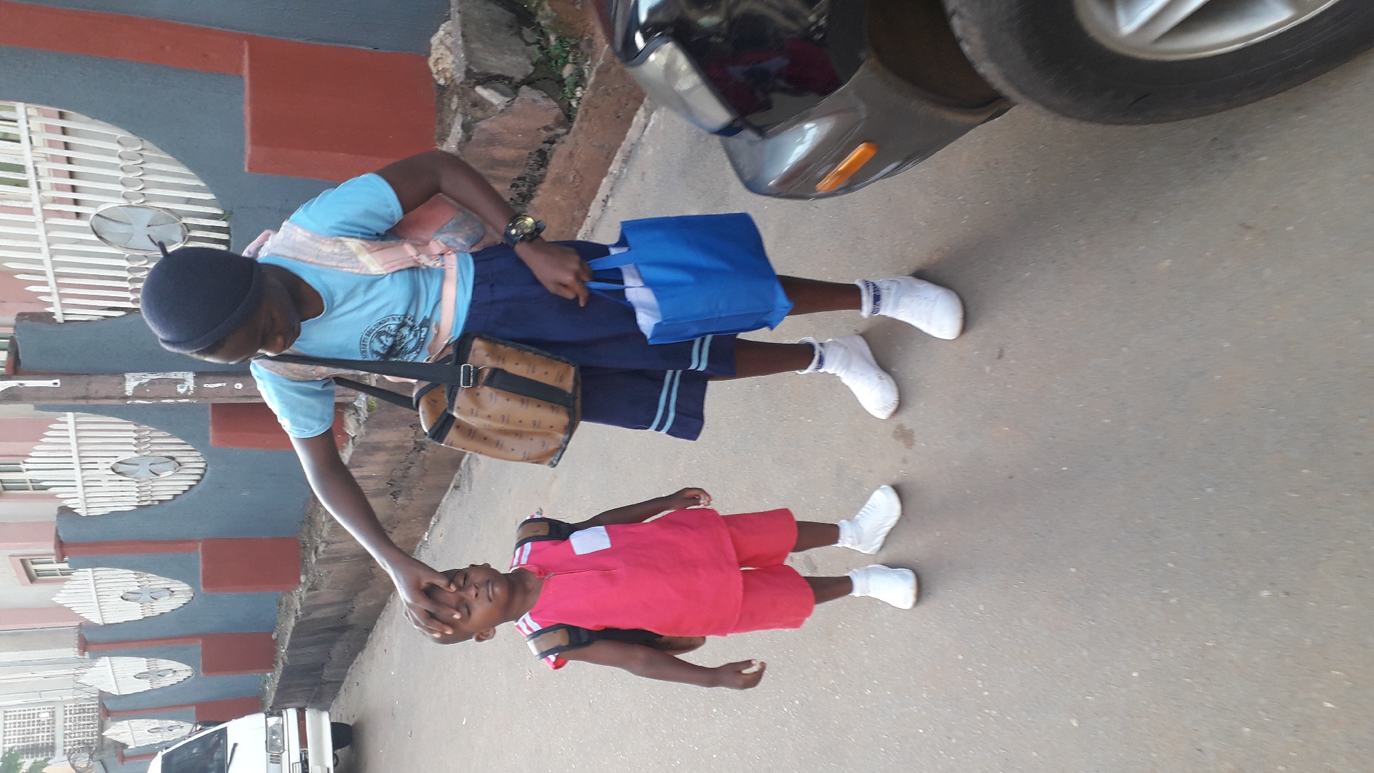 Two children are standing on an asphalt road next to a car, one of whom is being touched by the other. The older child wears a black beanie, a light blue t-shirt, a dark blue pair of shorts, white socks, and white shoes. They also have a brown bag and a blue tote bag with white handles and a white interior. The younger child is wearing a pink short sleeve shirt, pink shorts, white socks, and white shoes. They appear to have a small black backpack on. In the background, there is a building with grey and maroon sections and a white metal gate. Also visible are parked cars on the left. The picture appears to be taken in Onitsha, Nigeria.