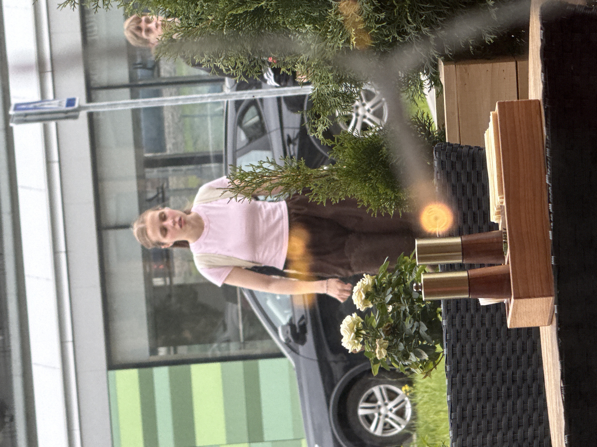 The image captures an outdoor scene, likely a cafe patio or terrace, in Moscow, Russia, during daytime. A young woman with light brown hair is the main subject, positioned in the lower left quadrant of the frame (when oriented correctly). She wears a light pink short-sleeved top and dark brown trousers or skirt, looking towards the viewer with a slight smile. A watch or bracelet is visible on her left wrist.
To her right, a black woven outdoor chair and a wooden table stand or shelf are visible. On the table, two wooden and brass salt or pepper grinders are neatly arranged next to a small potted plant displaying white or cream flowers.
In the mid-ground, behind the woman and the outdoor seating, a dark-colored car with distinct silver alloy wheels is parked. Further in the background, a modern building features large, reflective glass windows. A blurred reflection of another individual, appearing to be a person with blonde hair, is visible in these windows, partially obscured by green foliage. A segment of a building facade with vertical green and light green stripes is also visible.
Lush green, pine-like foliage extends into the frame from multiple directions, creating a natural, somewhat dappled effect. A prominent bright, circular orange-yellow light flare appears in the mid-right area, possibly a lens flare or a strong reflection. A blue and white street sign can be vaguely seen in the background reflections, but any text on it is illegible. The natural lighting suggests a clear day.