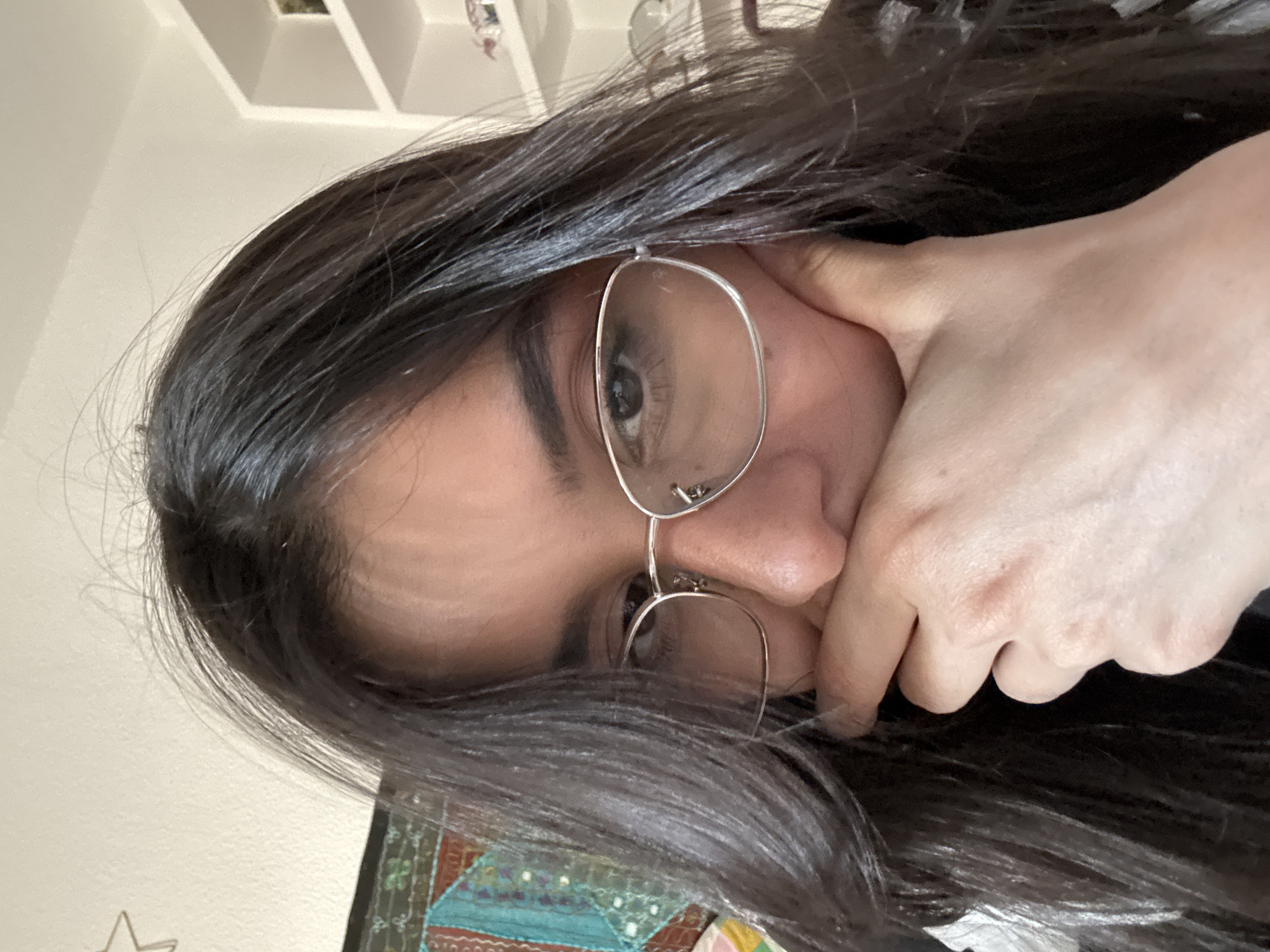 A close-up, low-angle selfie shows a young woman with long, dark hair and glasses looking towards the camera. Her right hand is raised, obscuring part of her mouth and chin, and her head is tilted slightly to the left. The background is out of focus but appears to be an indoor setting with a white wall and shelving unit visible. A portion of a colorful tapestry or textile is visible on the left side of the image.