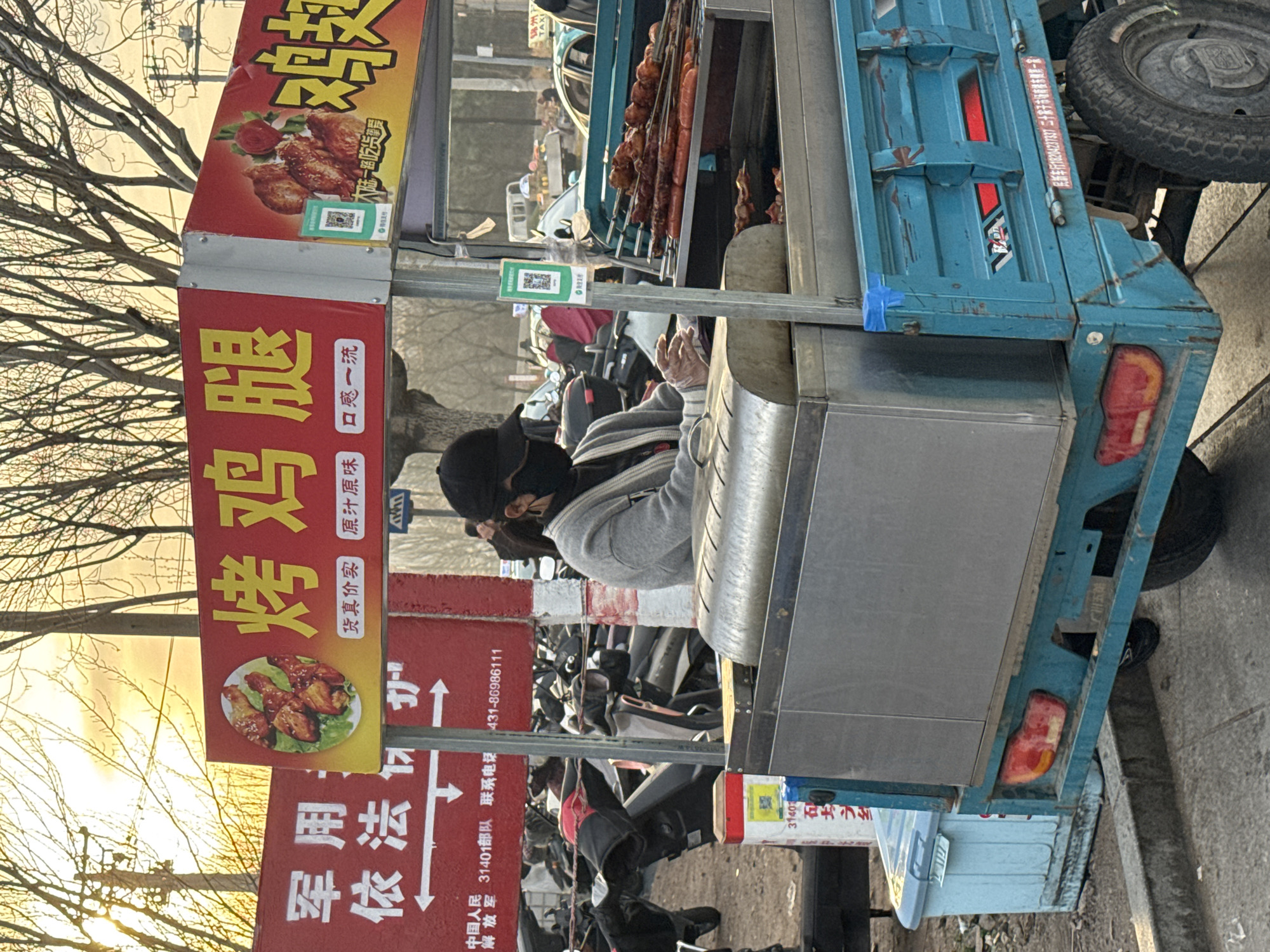 The image shows a street food vendor in Zhongtun, China, selling grilled chicken legs. The vendor is a person wearing a mask, a grey hoodie, and a black hat. The food cart is a blue, three-wheeled vehicle with signs in Chinese advertising the food. The environment includes a street with other parked vehicles and trees in the background. A red sign indicates the presence of the Chinese People's Liberation Army 31401 unit. The OCR text reveals the phone number to be 431-86986111.