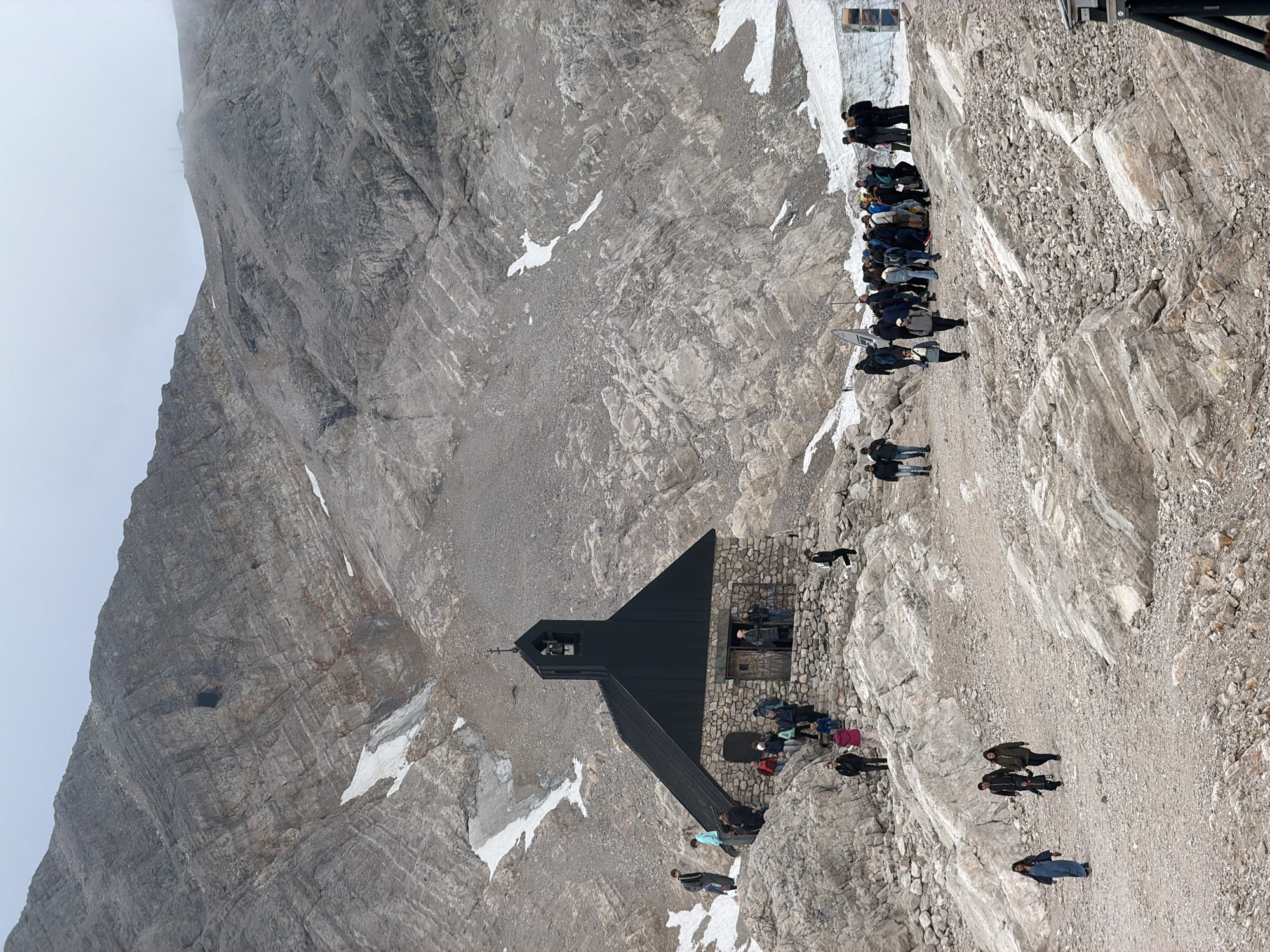 The image depicts a mountain landscape with a small stone church featuring a dark, pitched roof and a steeple with a bell. The church is situated on a rocky, uneven terrain with patches of snow. Several people are gathered around the church, some walking on the rocky paths and steps leading to the entrance, while others are standing in groups. The background is dominated by a large, rugged mountain slope composed of grey rock and scree. The sky is overcast and hazy. This scene is located at the Zugspitze summit in Garmisch-Partenkirchen, Germany.