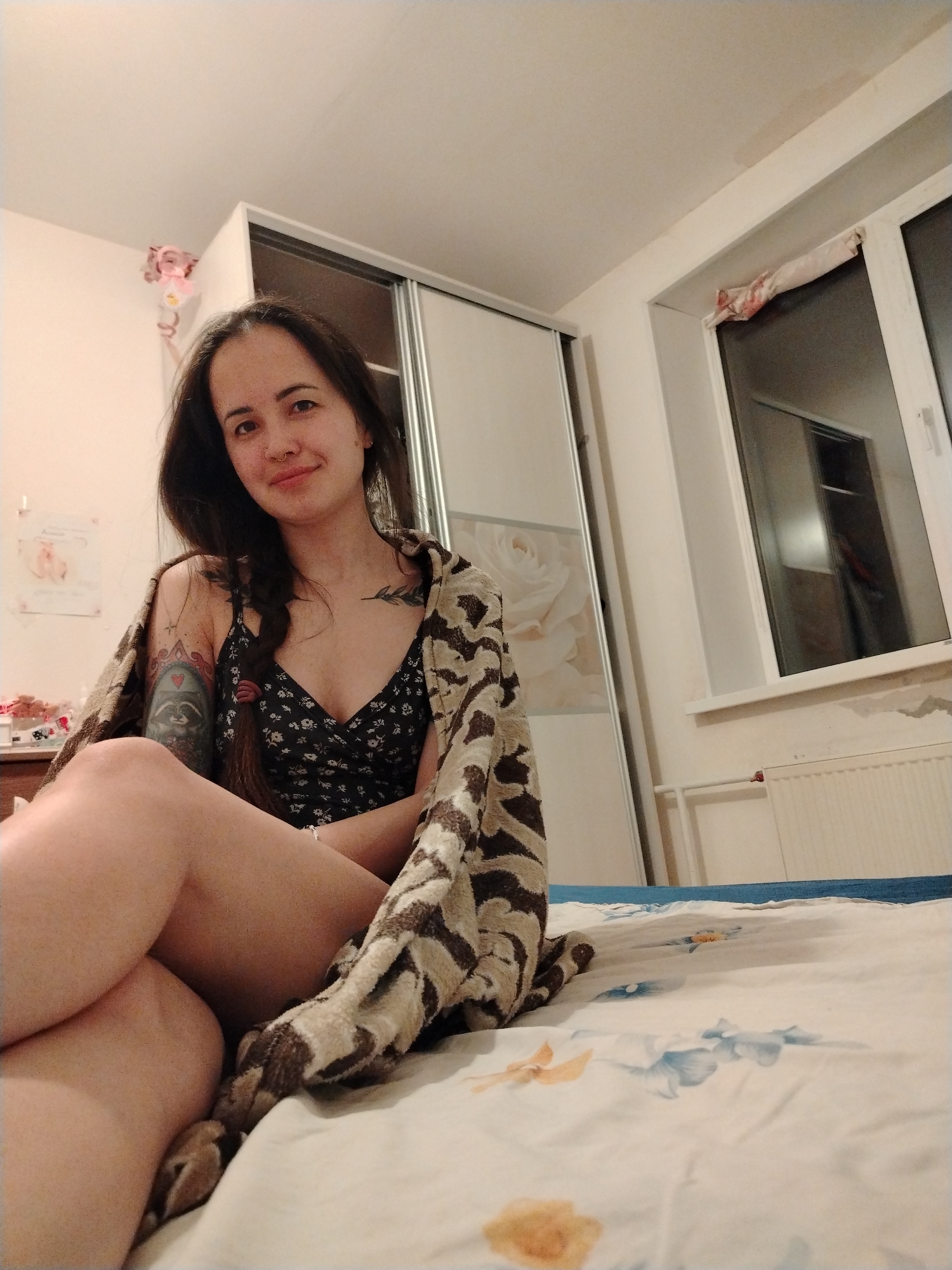 The image depicts a woman seated on a bed in a room. The woman has dark hair styled in a braid, fair skin, and tattoos on her arm and chest. She is wearing a floral-patterned tank top and has a patterned blanket draped over her shoulders. She is looking towards the camera with a slight smile.
The room appears to be a bedroom. There is a wardrobe with sliding doors, one featuring a floral design, against a wall. A window is visible with a radiator beneath it. The bed has a white and blue floral patterned bedsheet.
There is no indication that this image was taken in the city of Noviy, Russia.