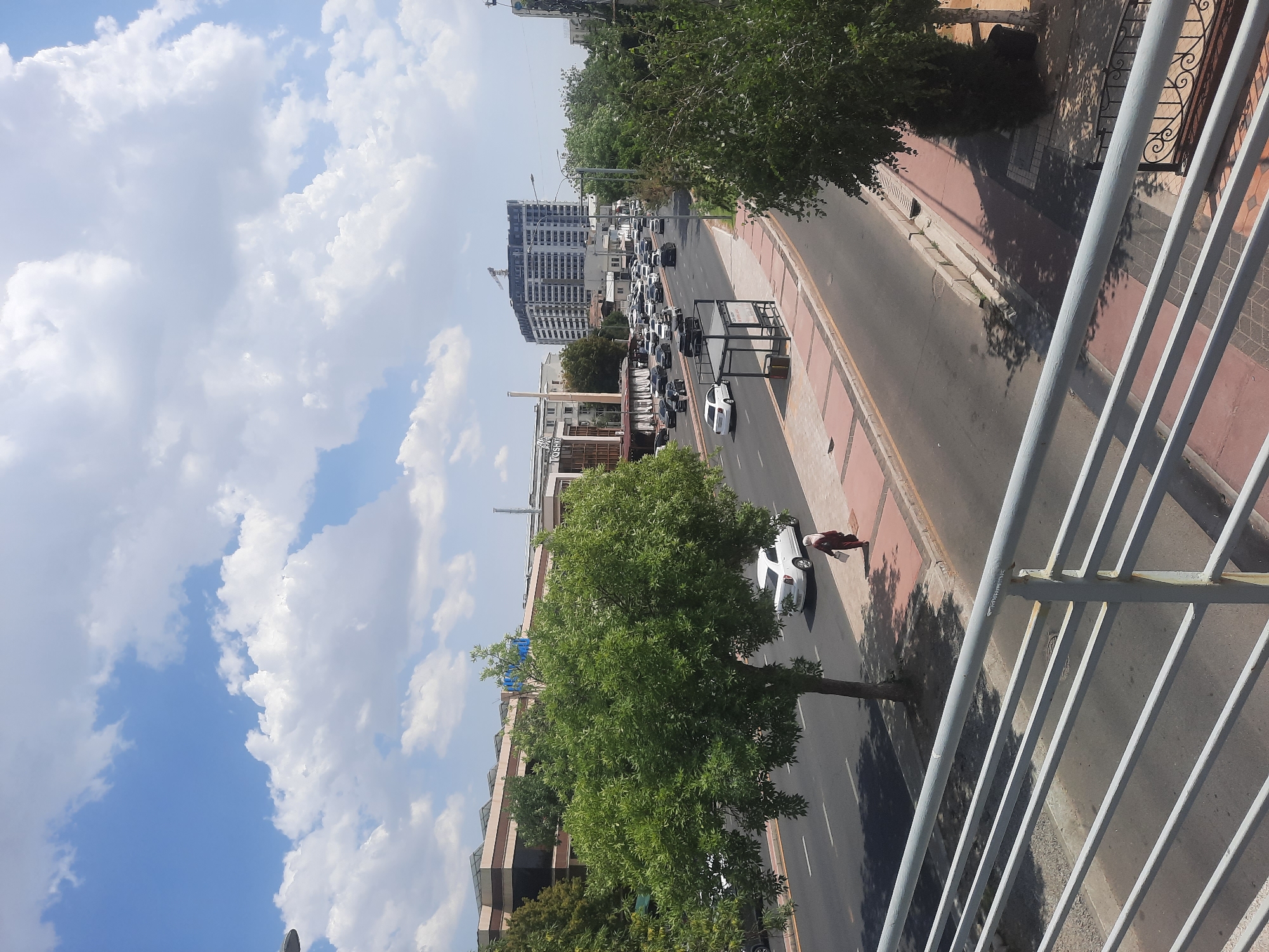 The image depicts a street scene in Tashkent, Uzbekistan, likely during daylight. A multi-lane road runs horizontally across the frame, with moderate traffic consisting of numerous cars. A white car is stopped on the right side of the road, and a woman wearing a dark headscarf and a maroon dress is walking on the sidewalk adjacent to it. On the left side of the road, a large, lush green tree dominates the foreground, casting a shadow on the asphalt. Beyond the tree, buildings with modern architecture are visible. Further down the road, a tall, contemporary building under construction is prominent, with a crane at its summit. The sky above is blue with scattered white clouds, indicating fair weather. A bus stop shelter with seating is situated on the right side of the road. In the foreground, a metal railing of a pedestrian overpass or bridge is visible, suggesting the image was taken from an elevated position. The sidewalks are paved with red and grey tiles.