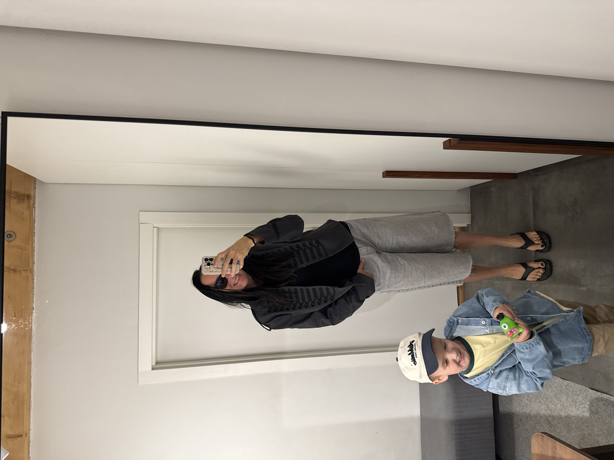 A woman is taking a mirror selfie in what appears to be a dressing room or fitting room. She is wearing a dark jacket with ornate detailing, a black shirt, light gray shorts, and black flip-flops. She is smiling and holding a smartphone. To her left, a young boy in a denim jacket, yellow t-shirt, and a baseball cap is holding a small green toy. The environment is sparse, with white walls and a wooden beam visible at the top. The reflection in the mirror shows the back of the room, which is also white and appears to be a long hallway or area. There is no indication that this scene is located in Chaikovsky, Russia.