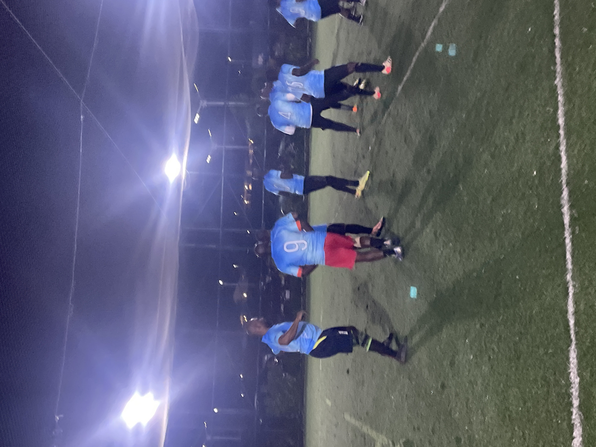 A group of men in athletic attire are gathered on a brightly lit artificial turf sports field at night, possibly for a game or practice. The field is marked with white lines and appears to be enclosed by a net or fence overhead. Several men are wearing blue jerseys with different numbers visible on their backs, including "4," "6," and "9." Some are wearing black shorts, while others have red or blue shorts. One man in the foreground is wearing a blue jersey, black shorts, and green and black striped leg coverings. The lighting appears to be artificial stadium lights, creating a somewhat hazy atmosphere with lens flare. The background shows some indistinct structures and lights, suggesting an urban or suburban environment. The overall scene depicts an organized sports activity taking place in a dedicated facility.