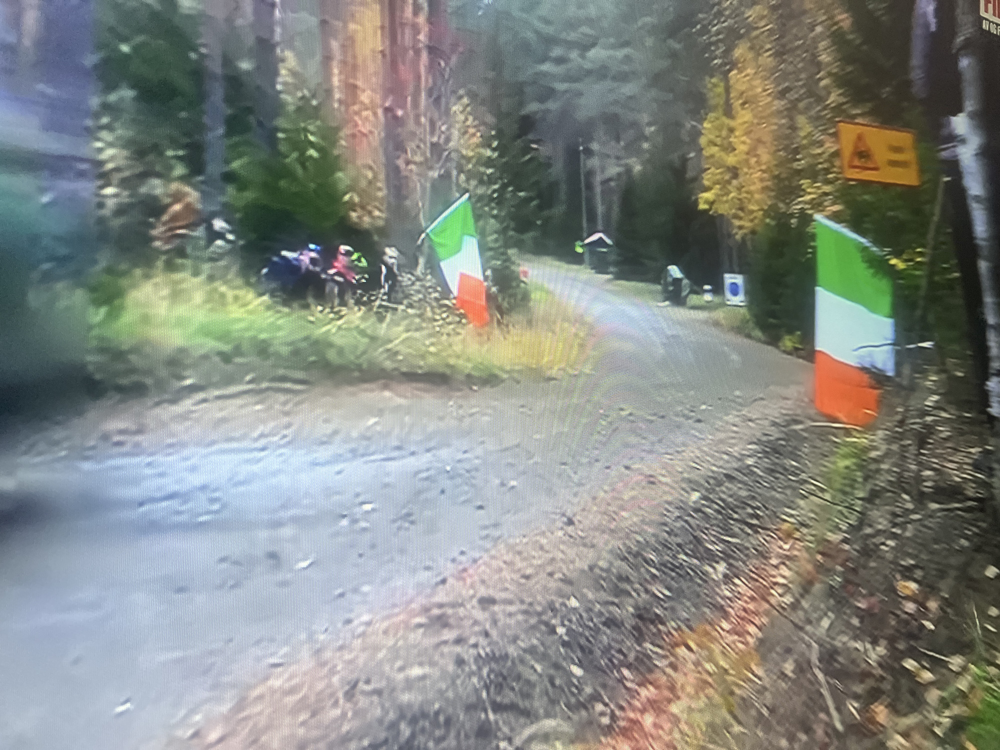 An outdoor, slightly blurry, medium shot shows a dirt road winding through a forest with autumnal trees. Two Italian flags are visible. One flag is on the left side of the road, and another is on the right. In the background, several people on motorcycles or ATVs are gathered on the left side of the road. A yellow warning sign is posted on the right side of the road in the background. The ground is covered in dirt and fallen leaves. The scene appears to be part of a rally or sporting event. The location is not identifiable as Masazir, Azerbaijan.