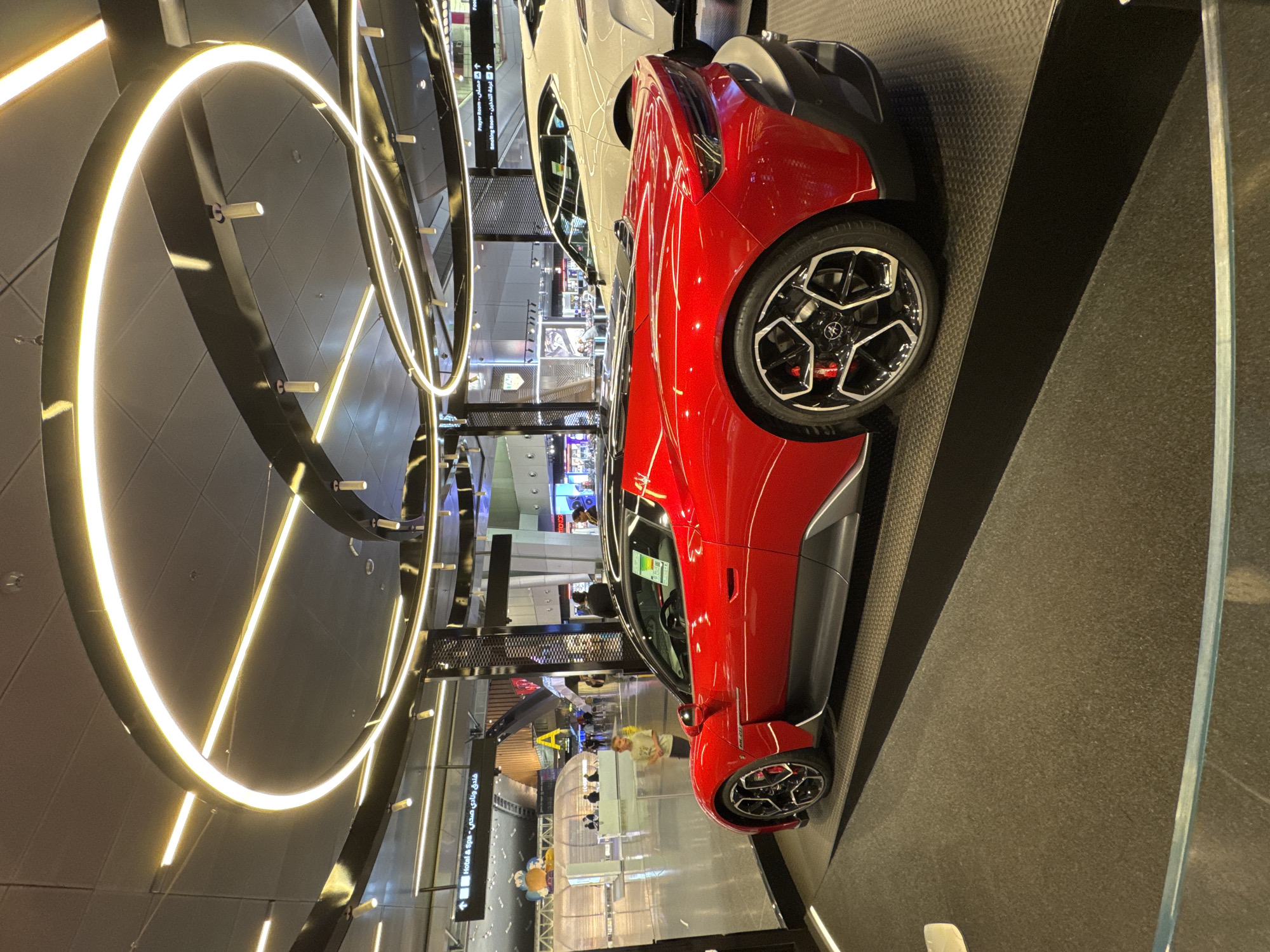 A bright red sports car, likely a Maserati MC20, is displayed on a black platform in what appears to be a modern airport terminal. The car is positioned in the foreground, viewed from its rear-three-quarter angle. It features a sleek design with aerodynamic lines, a low profile, and large, intricately designed black and silver wheels with red brake calipers visible.
Behind the sports car, a white SUV is partially visible, also likely on display. In the background, the airport environment is discernible with signage indicating directions to "Hotel & Spa" and "Prayer Room" in both English and Arabic. People are seen walking in the distance, some interacting with information desks or exhibits. The ceiling of the terminal features a striking modern lighting installation with concentric circular and linear fluorescent tubes emitting a warm yellow glow. The overall environment suggests a high-end exhibition or display area within a major transportation hub, possibly Hamad International Airport in Doha, Qatar, given the Arabic signage. A young child is standing to the left of the red car, looking towards the camera.